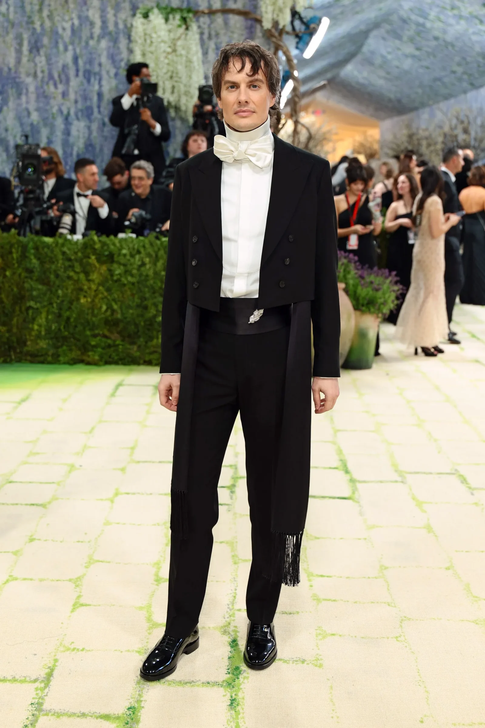 Man in a black tailcoat and white bow tie on a patterned carpet at a gala, with photographers in the background.