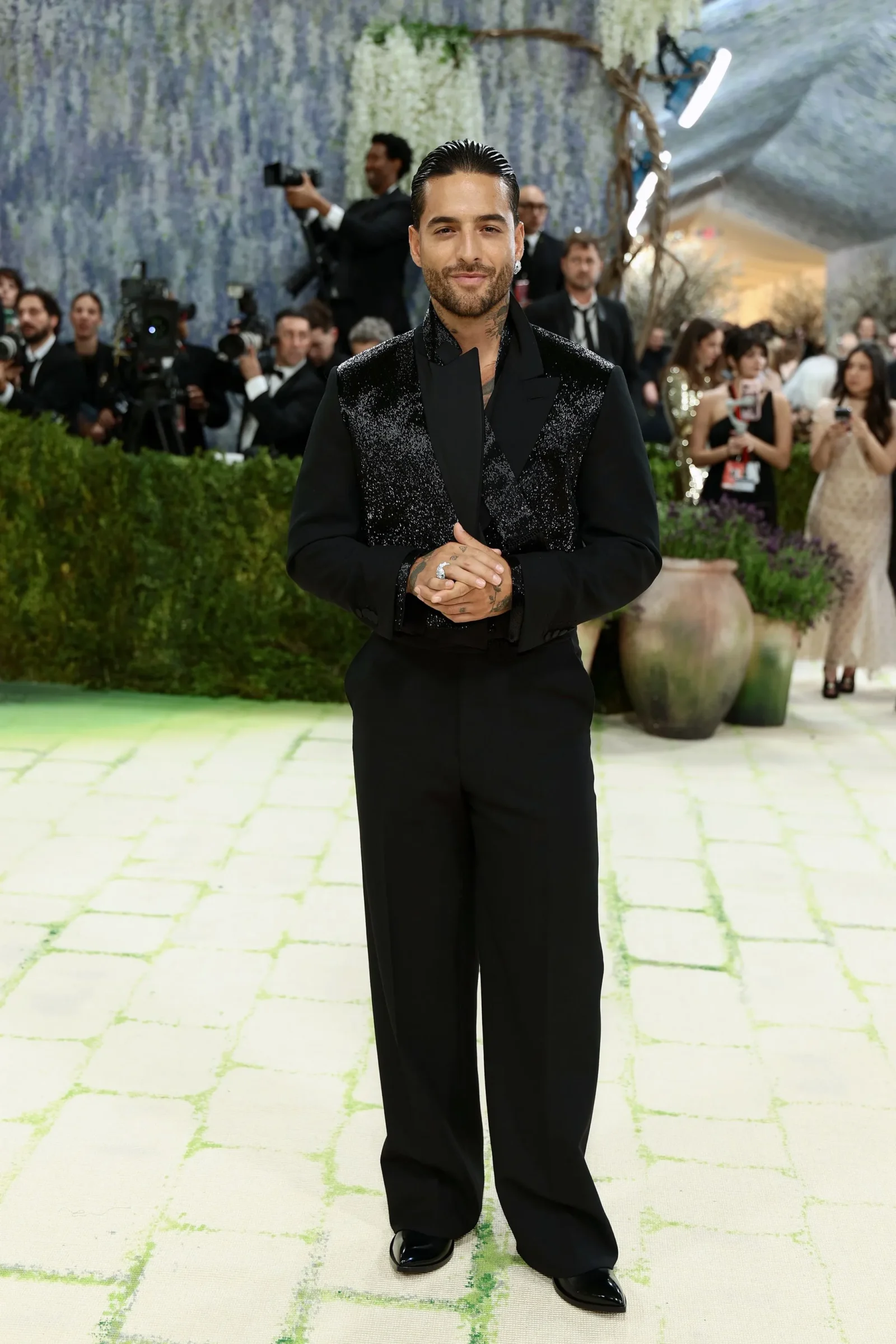 Man in a black glittered suit poses for cameras at a formal event, with photographers and guests in the background.