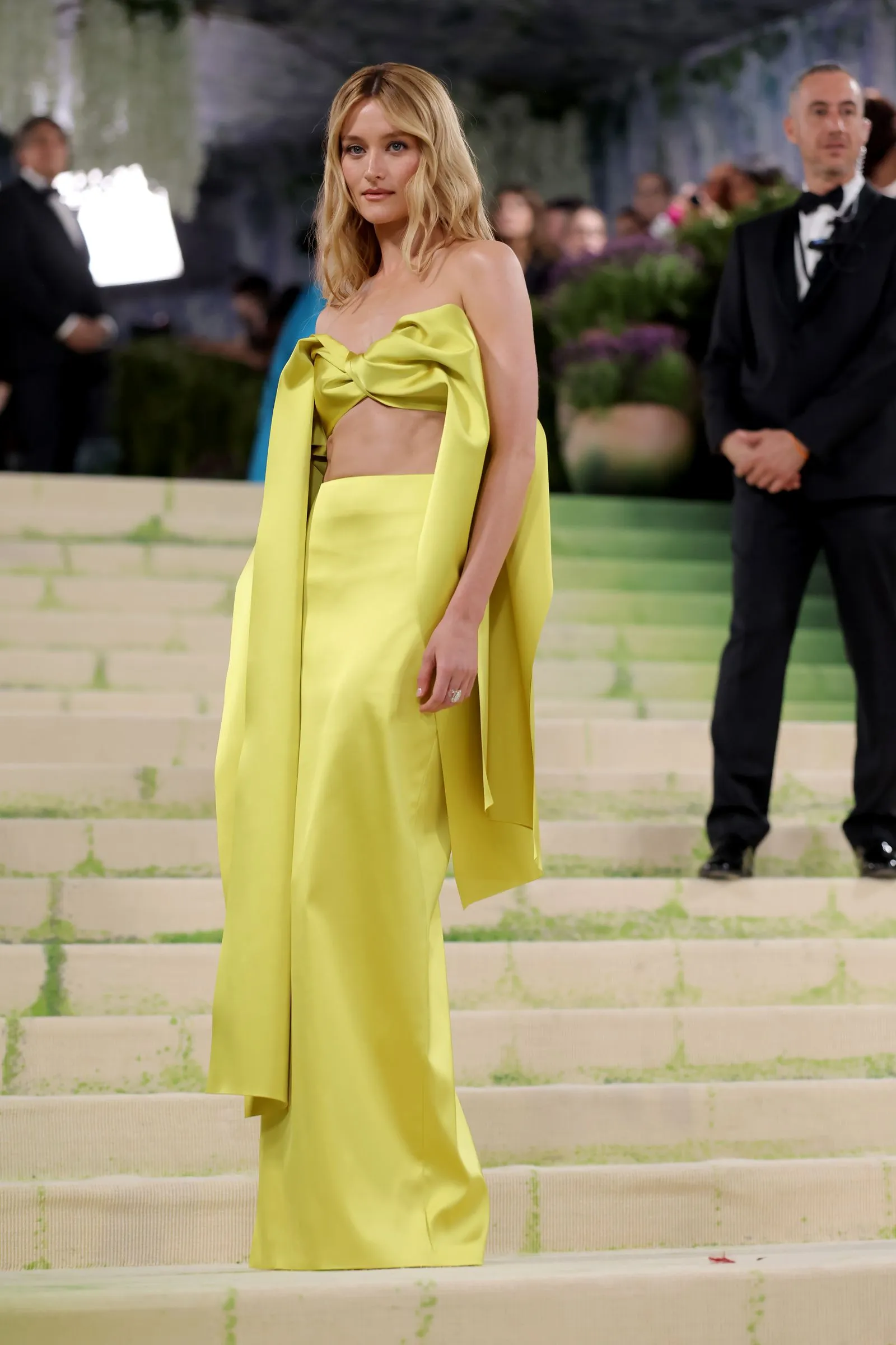 Blonde woman in a neon yellow two-piece outfit with a large bow, standing on light steps at a formal event.