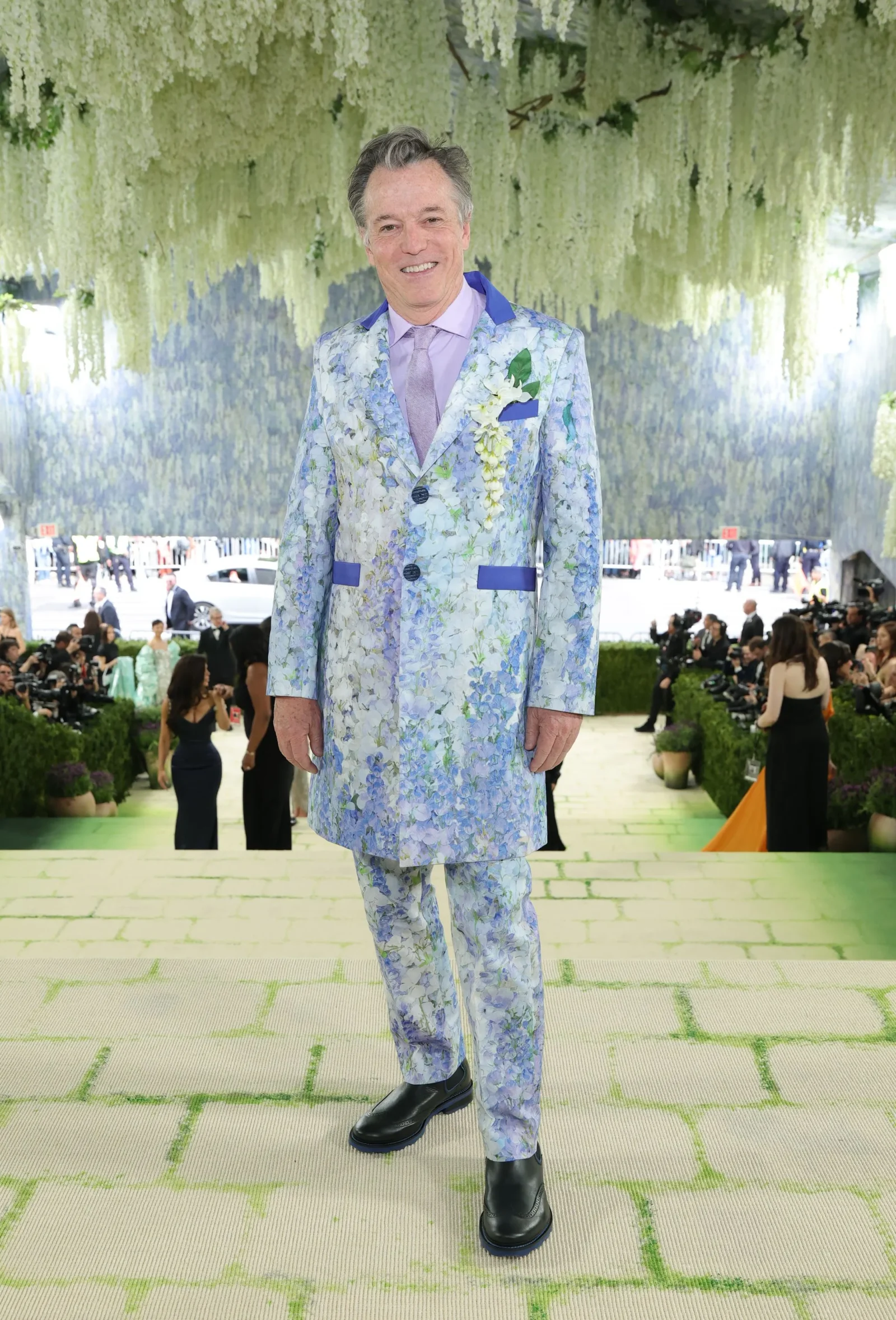 Man smiling in a blue floral suit with a lavender shirt at a fashion event, floral backdrop overhead.