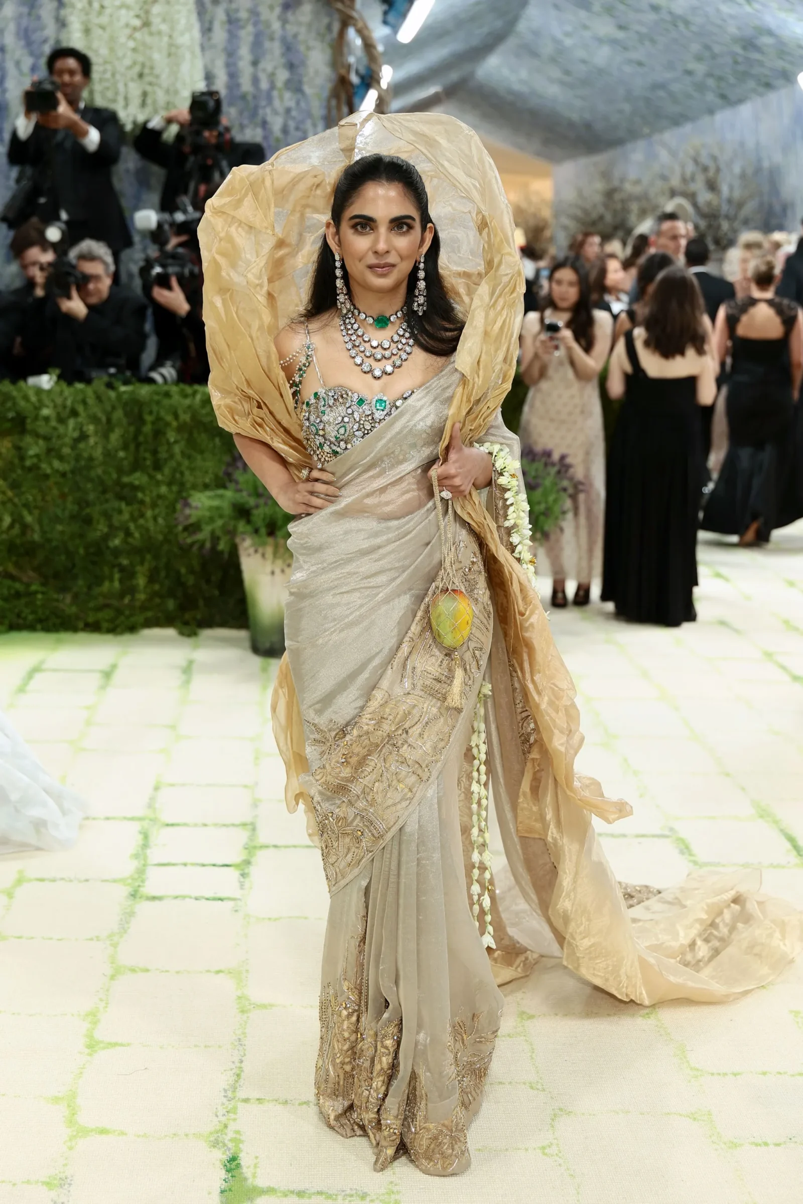 Elegant woman in a beige-gold sari with heavy diamond jewelry posing on a gala step-and-repeat, photographers in the background.