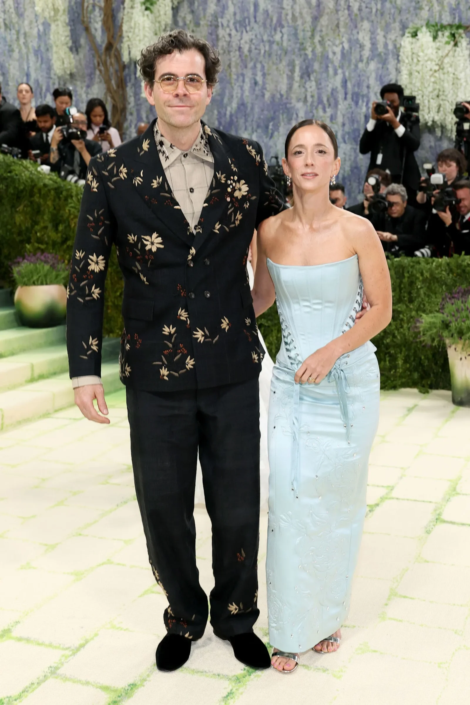 Man in a dark floral suit and a woman in a light blue gown pose together on a red carpet with photographers in the background, smiling softly to camera.