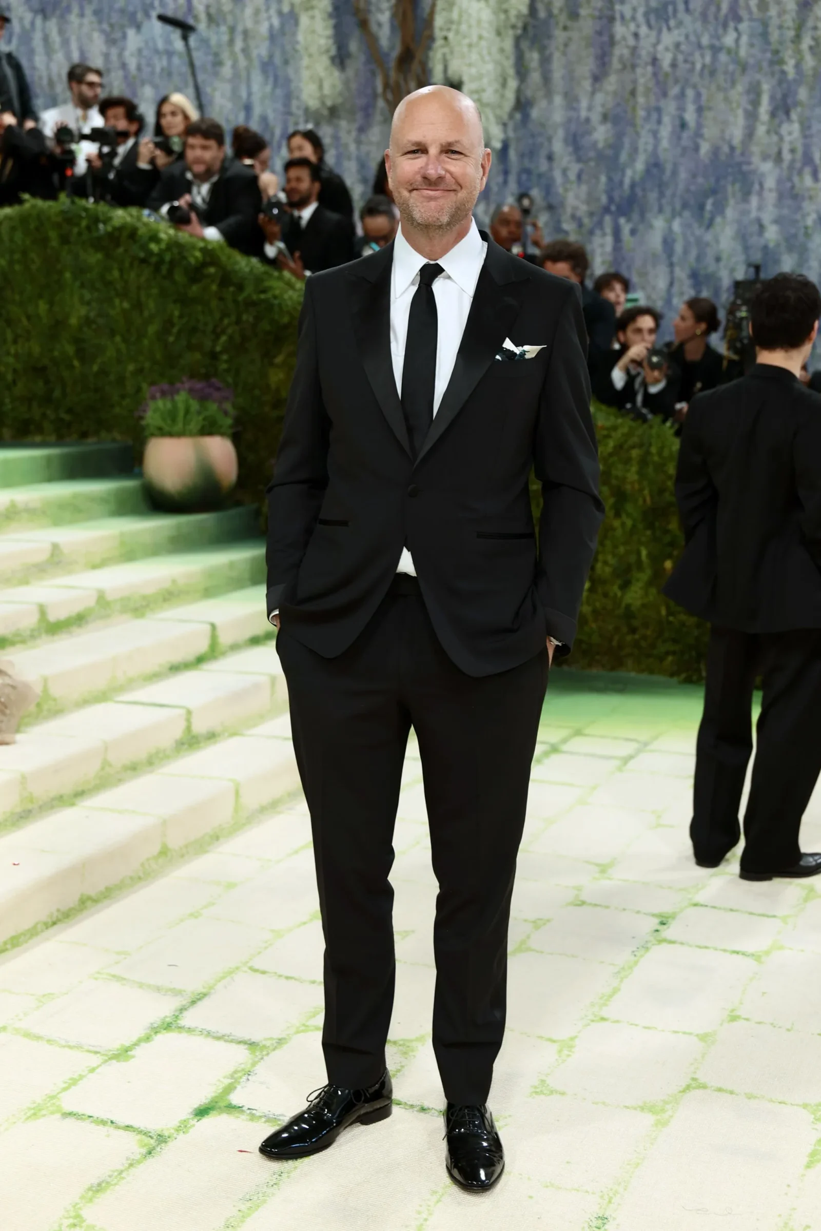 Man in a black suit and tie posing on light-green steps at a formal event, photographers in the background.