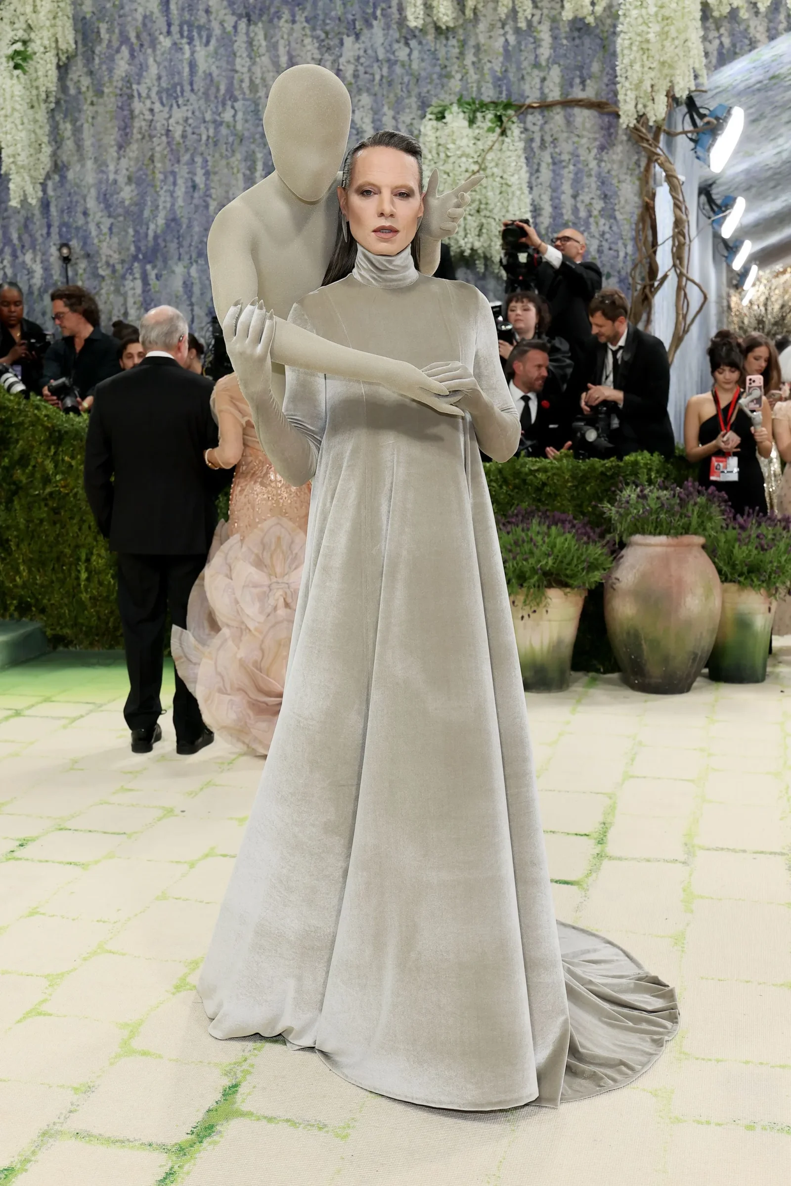 Model in a silver-gray, floor-length gown with oversized sculptural sleeves; a mannequin-like figure embraces her as photographers look on at a fashion event.