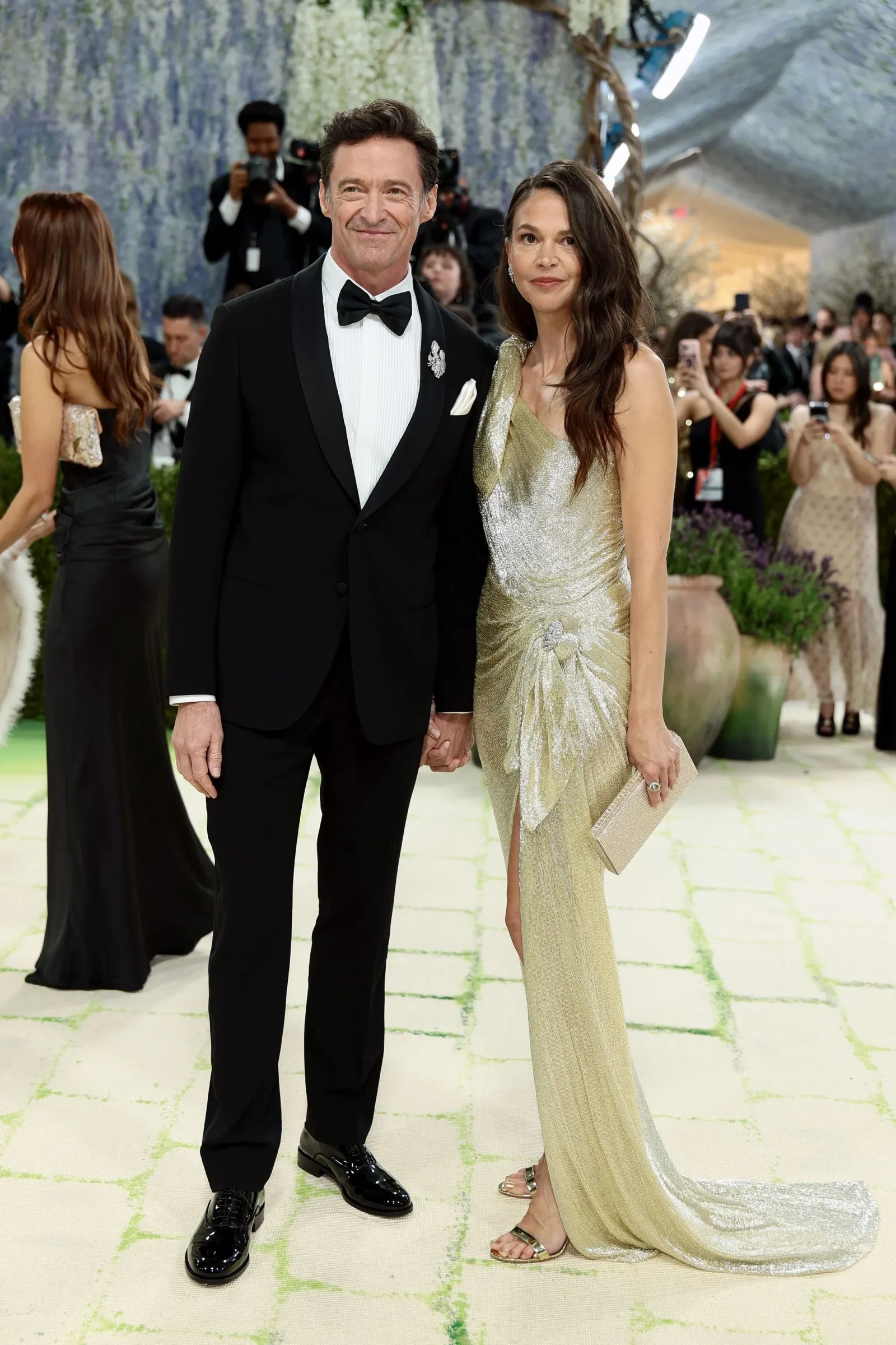 Smiling couple posing on a red carpet: man in a black tuxedo with bow tie and a jeweled lapel pin, woman in a shimmering gold gown holding a beige clutch.