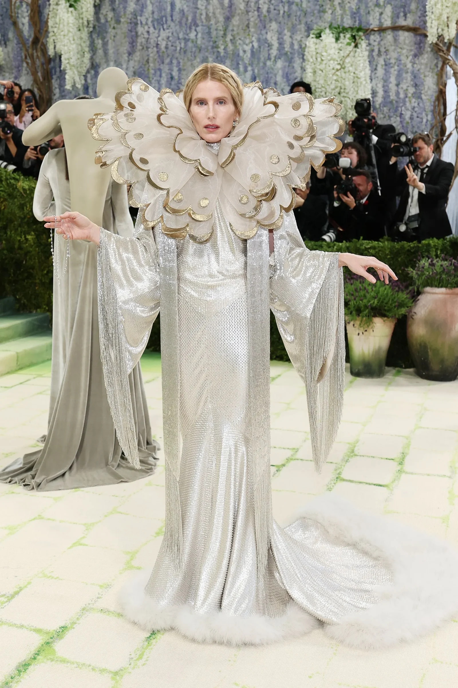 Person wearing a silver sequined gown with a large, scalloped gold-edged ruffled collar, posing on a fashion event run‑way with photographers in the background.