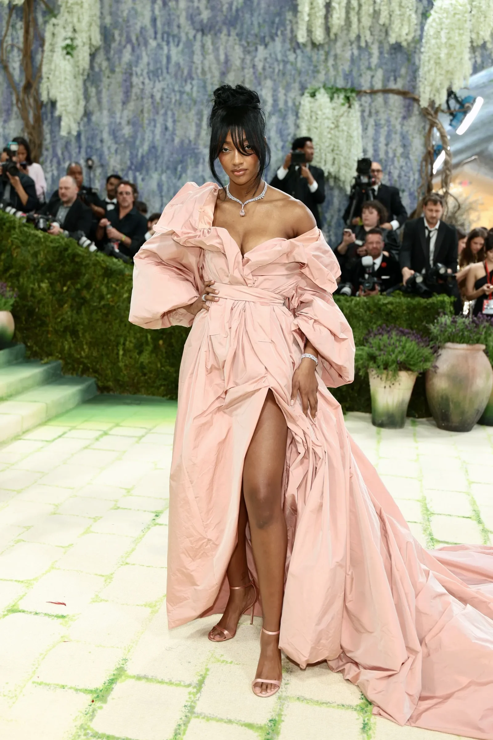 Woman in a voluminous pink gown posing on a green-tiled carpet at a formal event, surrounded by photographers and a floral backdrop.