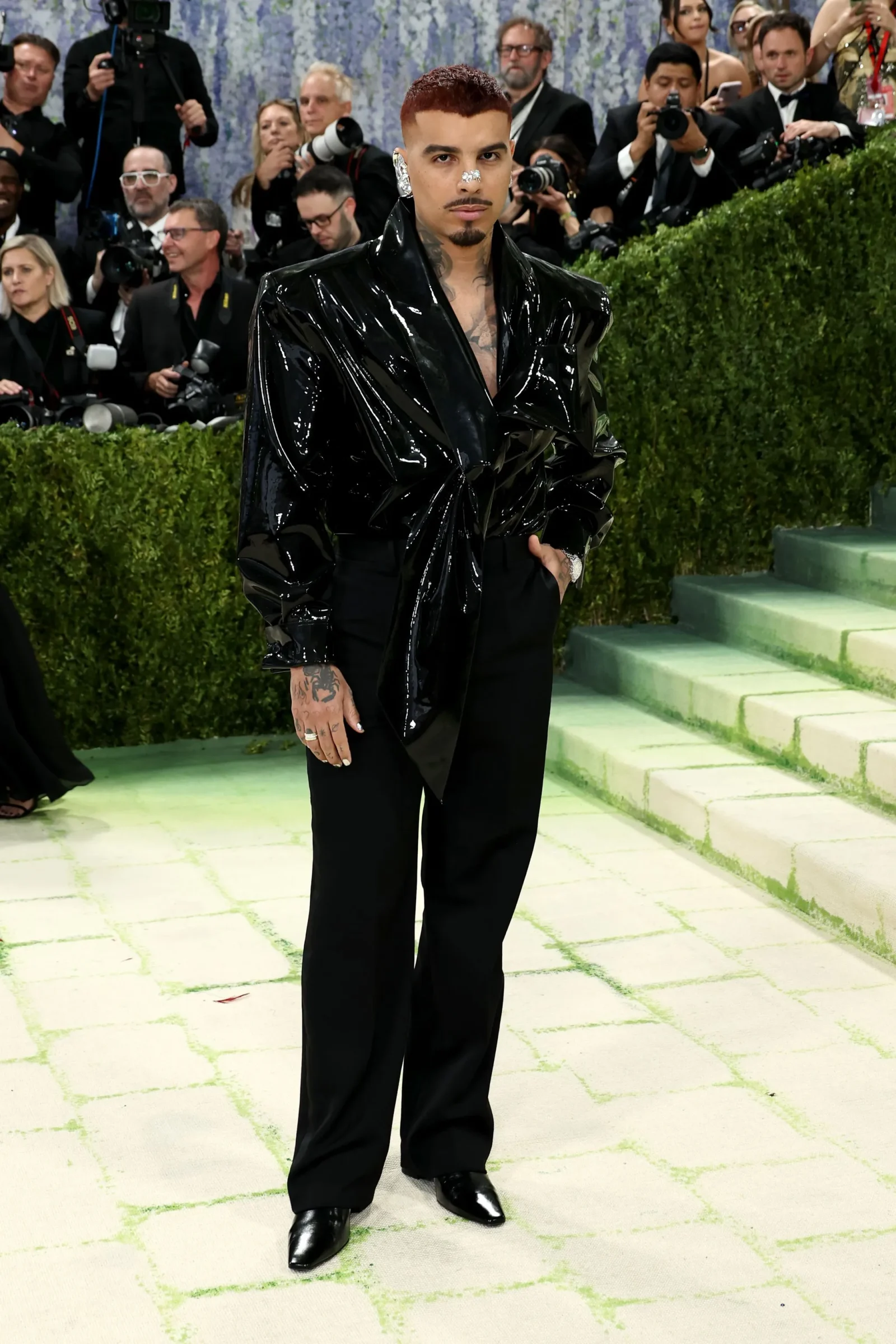 Man at a red carpet wearing a glossy black wrap coat and black pants, surrounded by photographers.