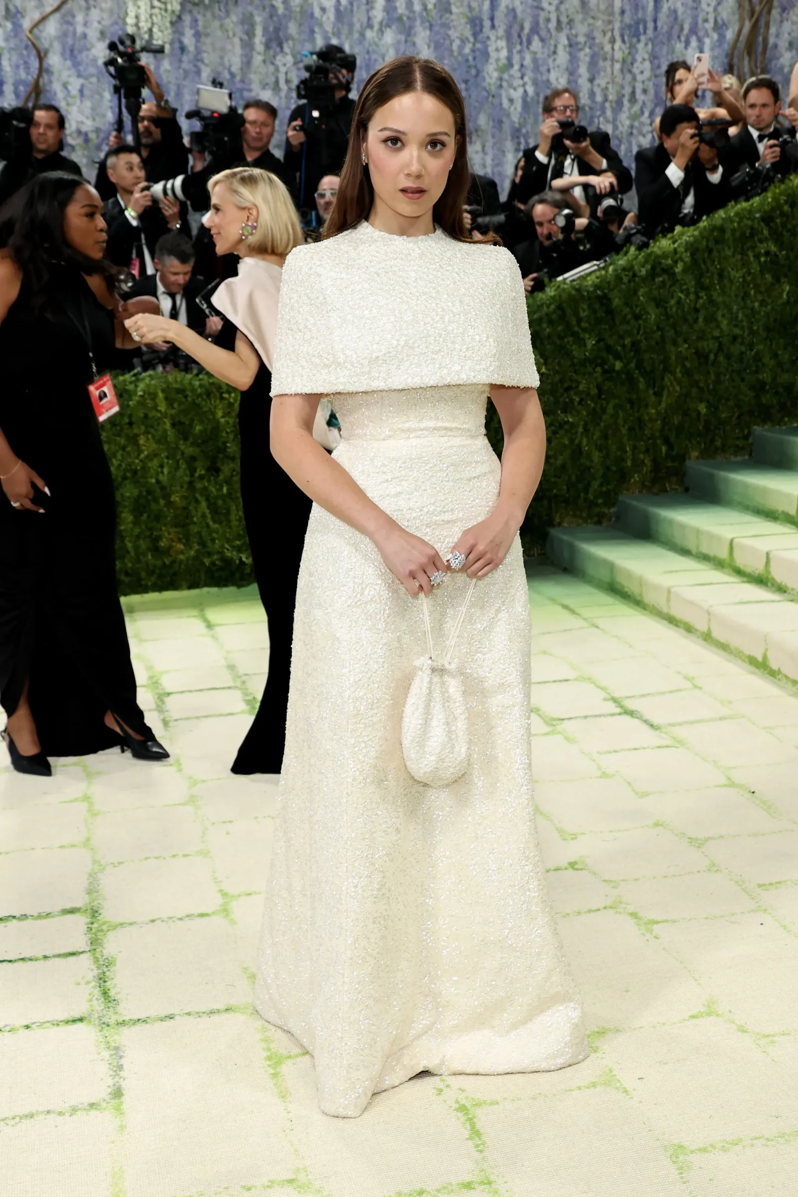 Woman in a white sequin gown posing on a green-tiled floor, hands clasped around a small purse, paparazzi in the background.