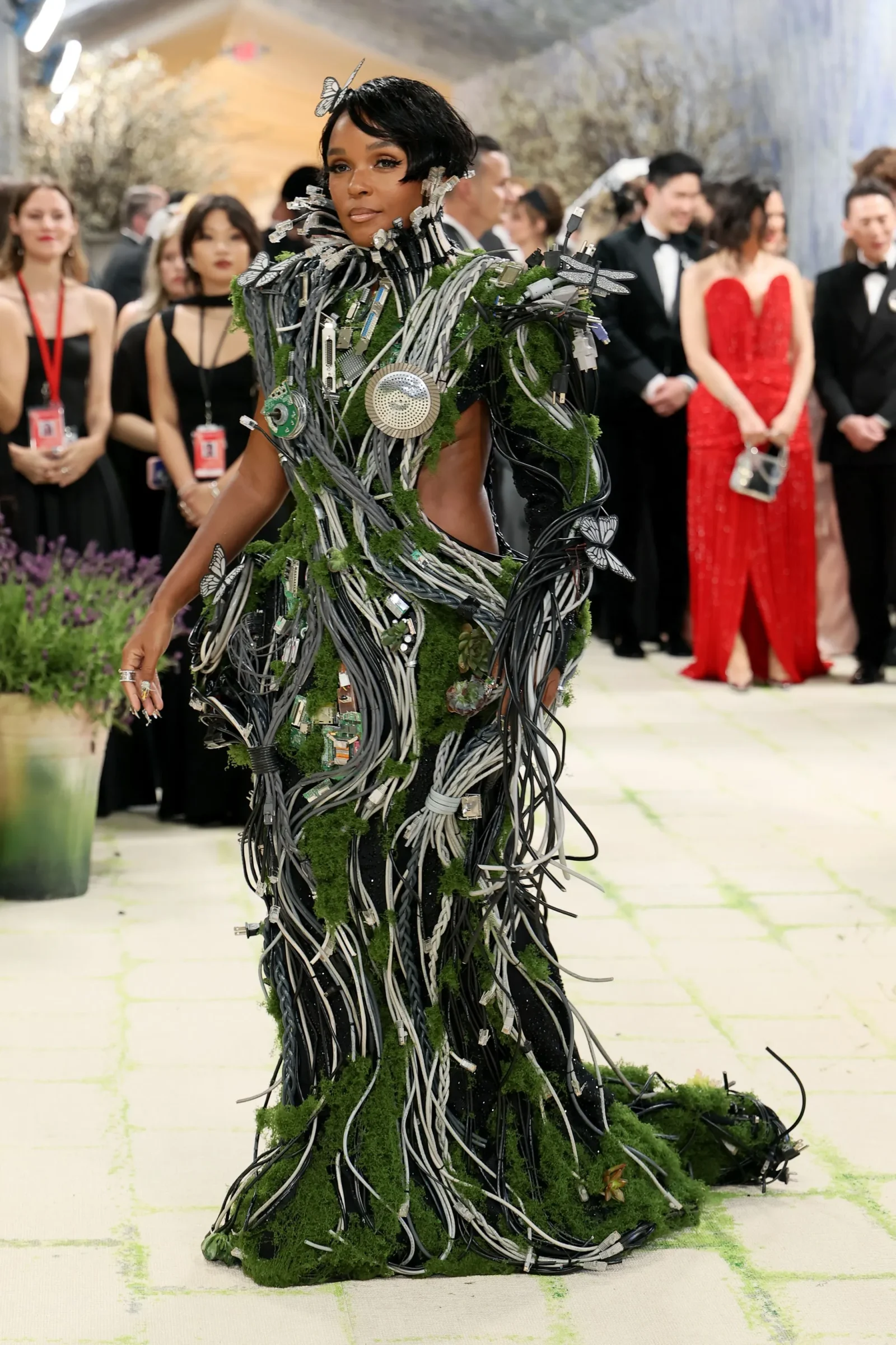 Model on a runway in a moss-covered, wire-sculpted gown with butterfly decorations and cables cascading down the outfit.