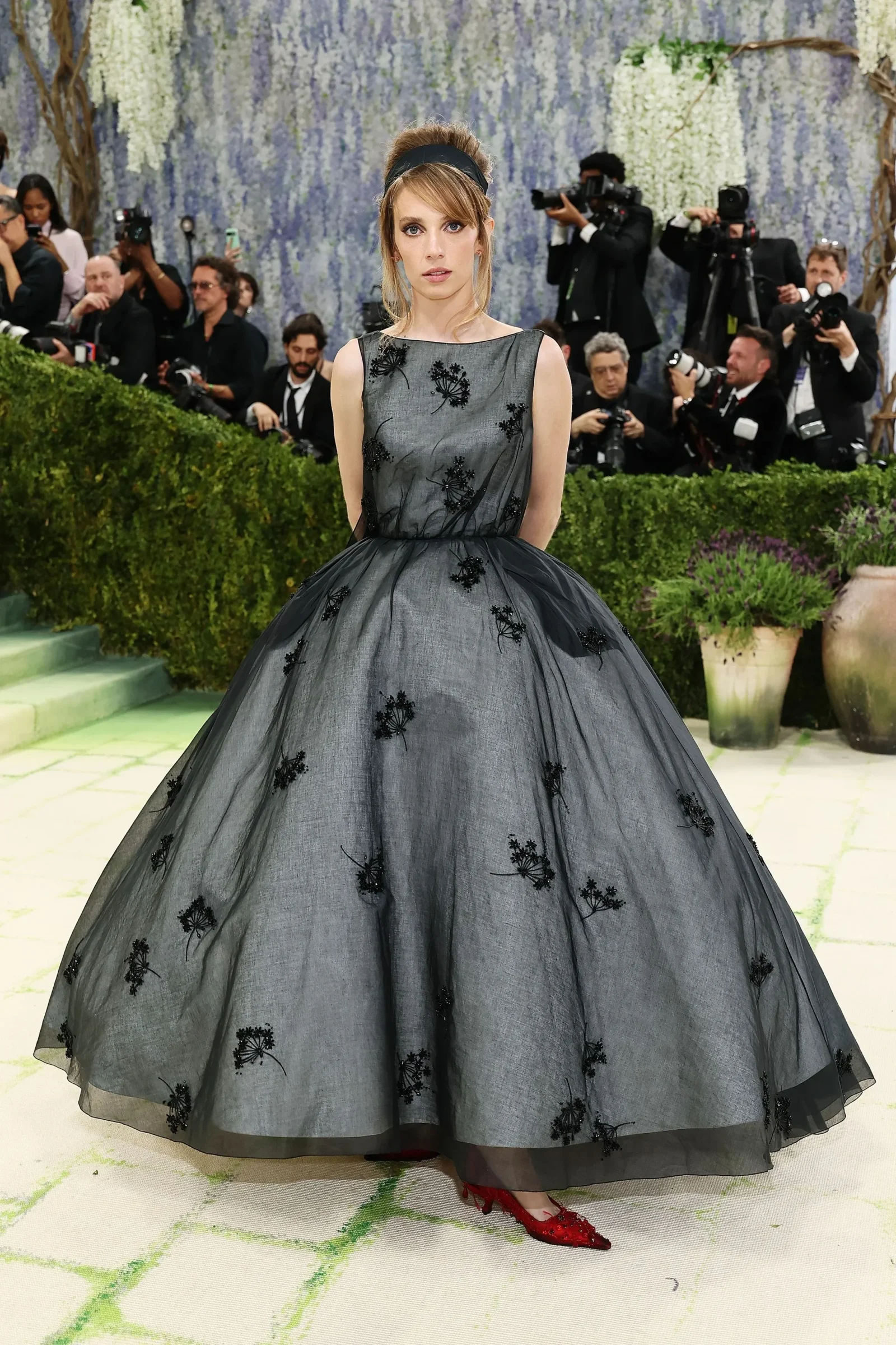 Woman in a voluminous gray gown with black floral embroidery poses on a carpeted step, surrounded by photographers and greenery.