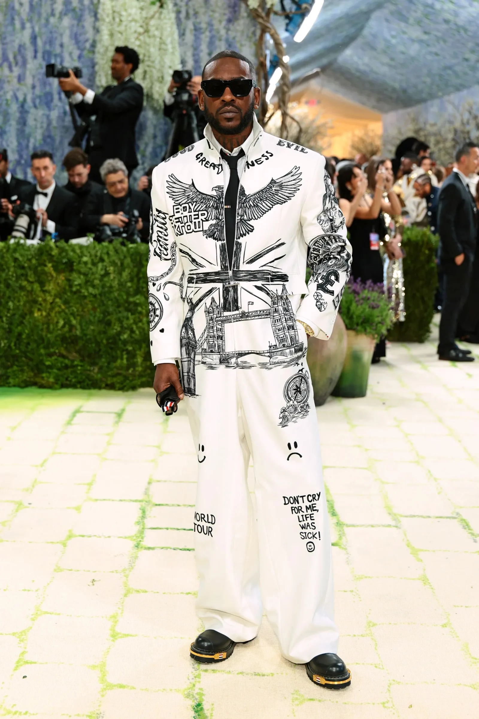 Man in a white graphic suit with wings and landmarks, wearing black sunglasses at a red-carpet event with photographers in the background.