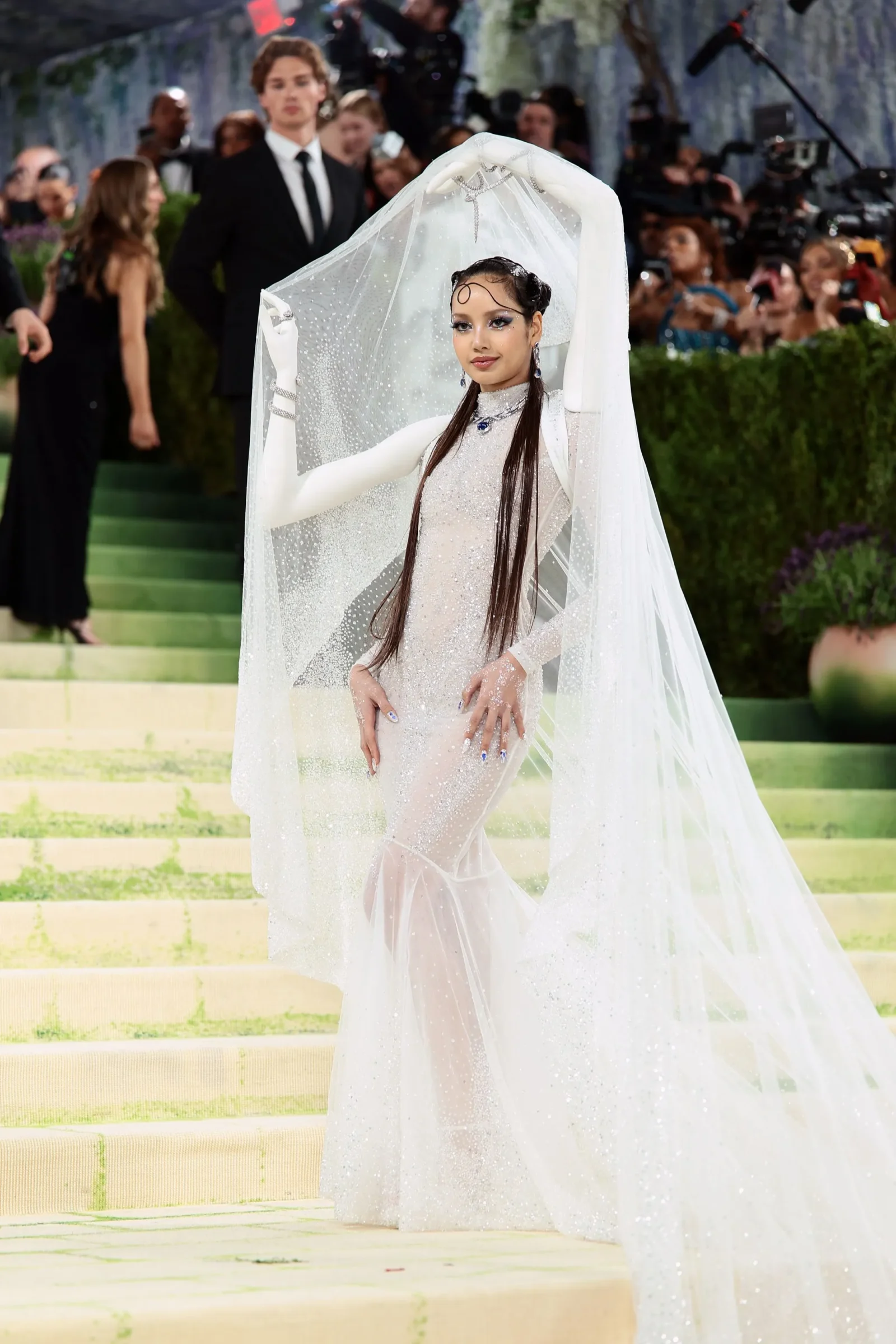 Rihanna posing on the red carpet in a sheer white beaded gown with a long veil, photographers in the background.