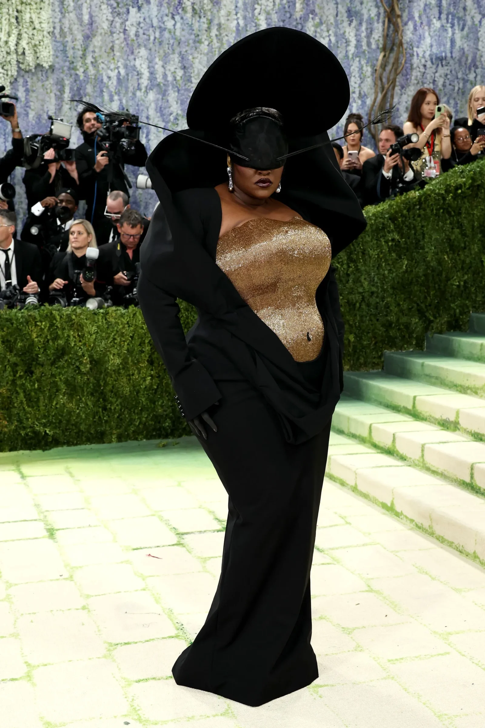Woman posing on a green carpet in a black suit with an enormous black hat and gold sequin top, surrounded by photographers.