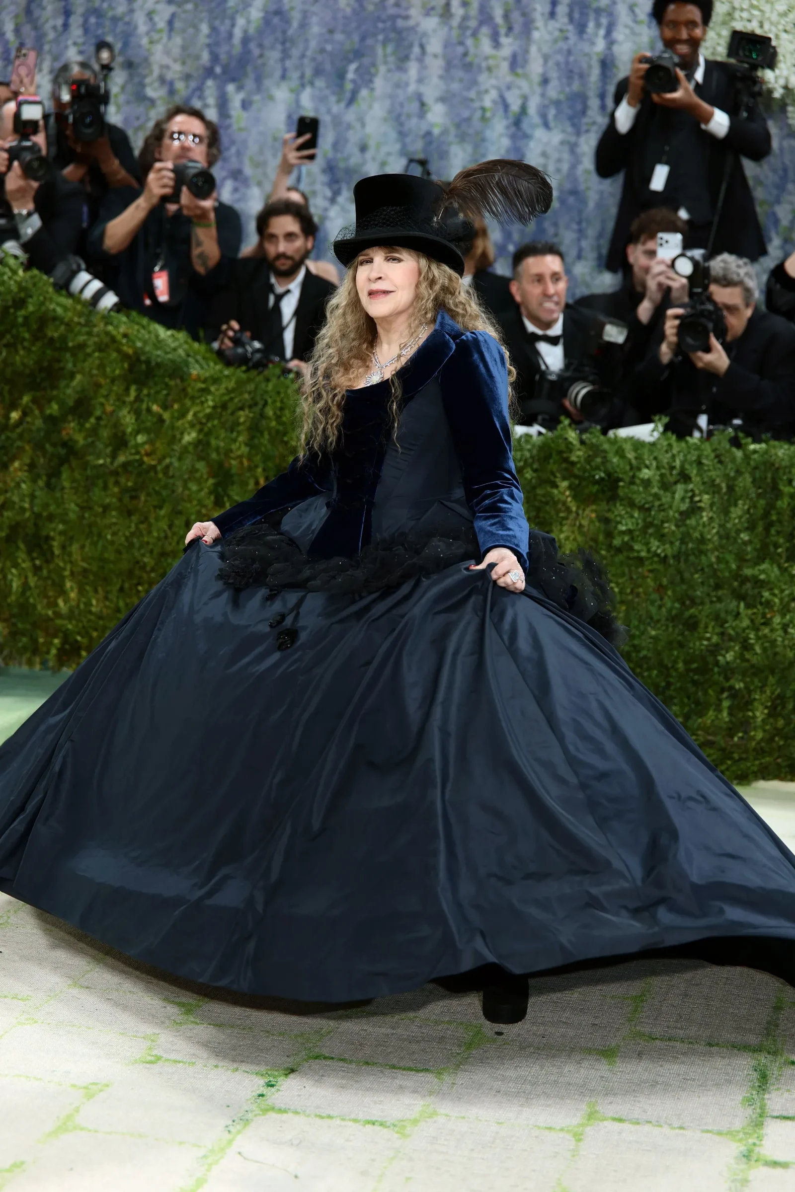 Woman in a dark blue velvet ball gown with a black feathered hat, posing for photographers at a gala.