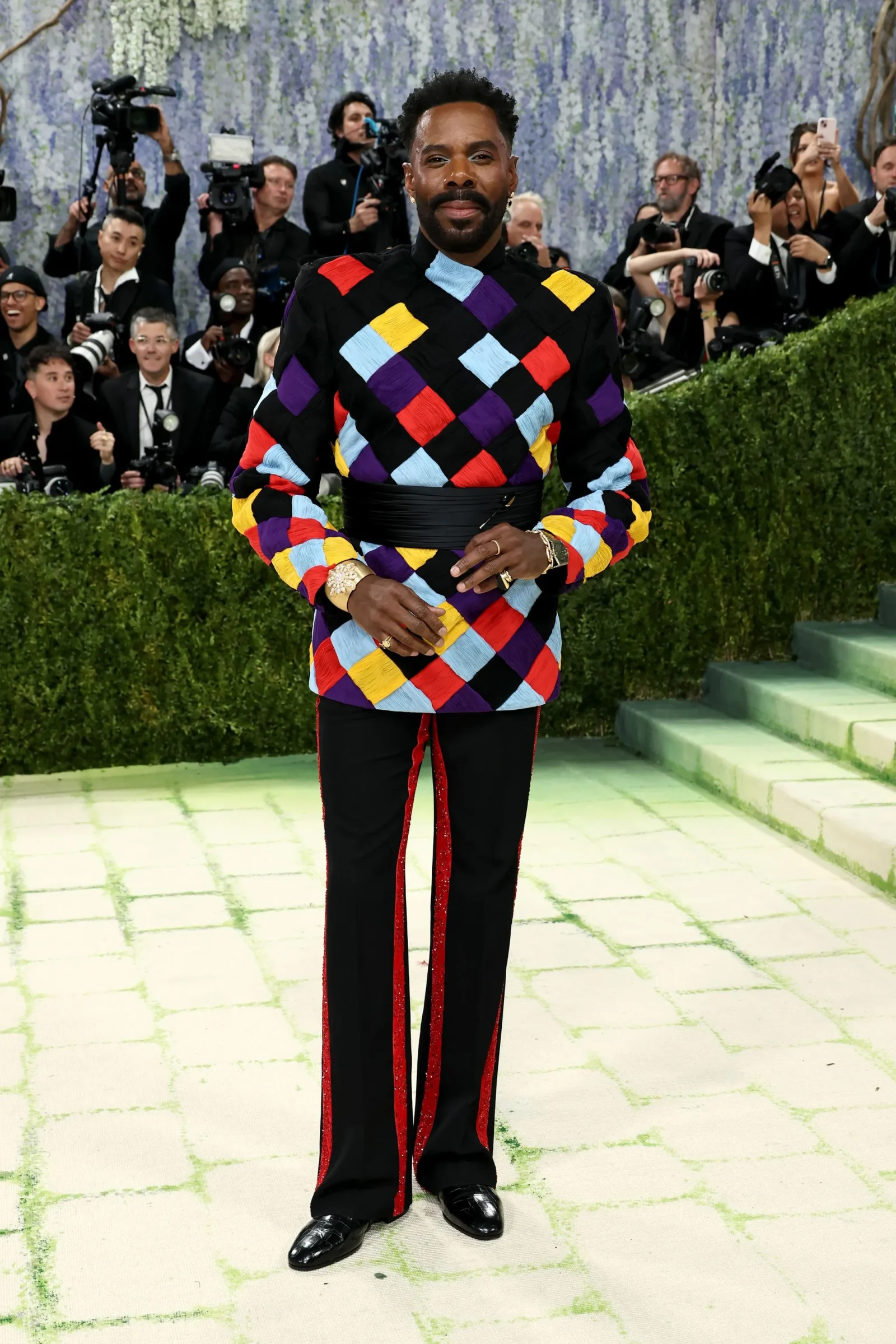 Man posing on a light tiled floor wearing a multicolored patchwork top and black pants with red trim, photographers behind him at a red carpet event.