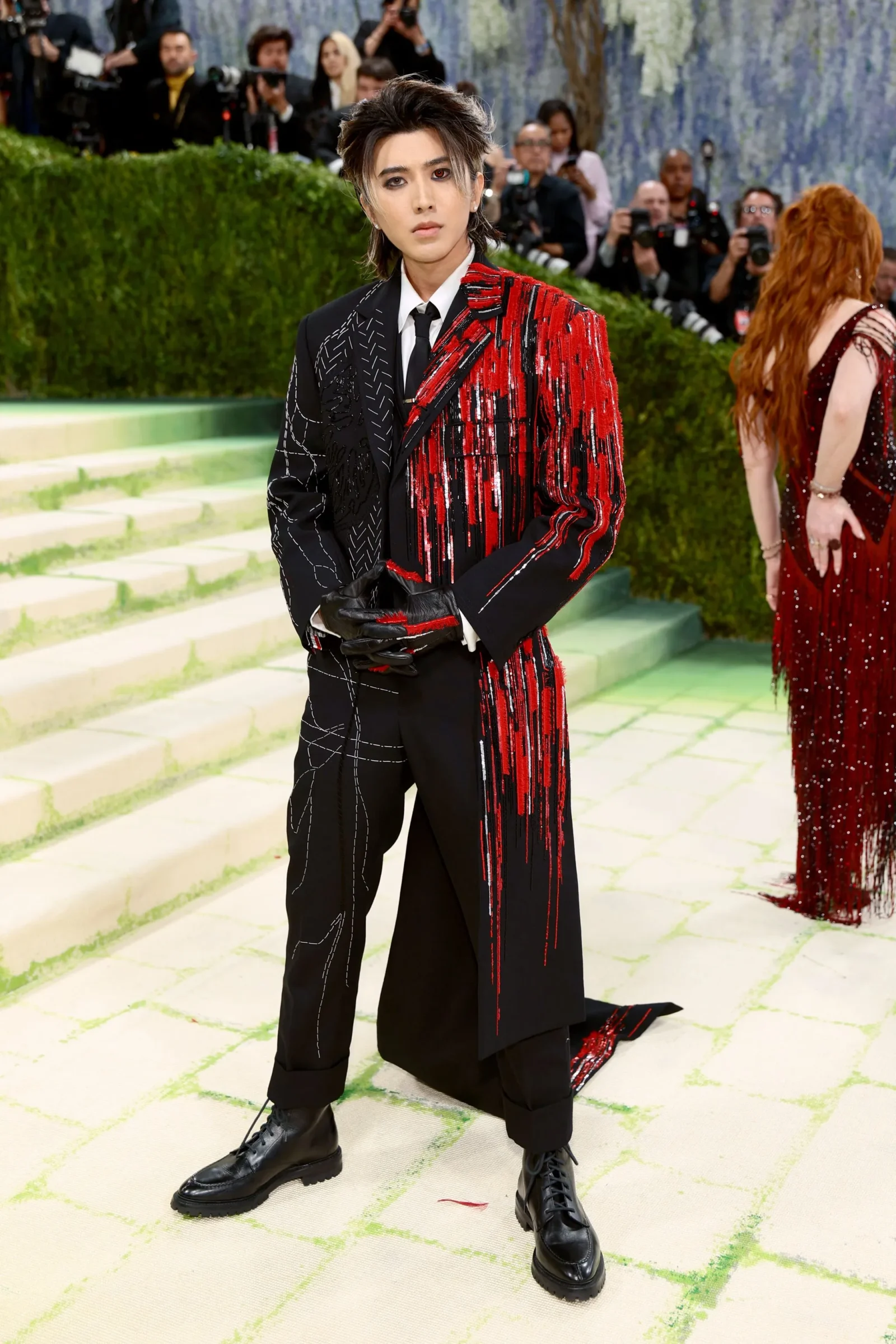 Person wearing a black-and-red sequined suit with gloves, standing on a pale green carpet at a red-carpet event as photographers watch in the background.