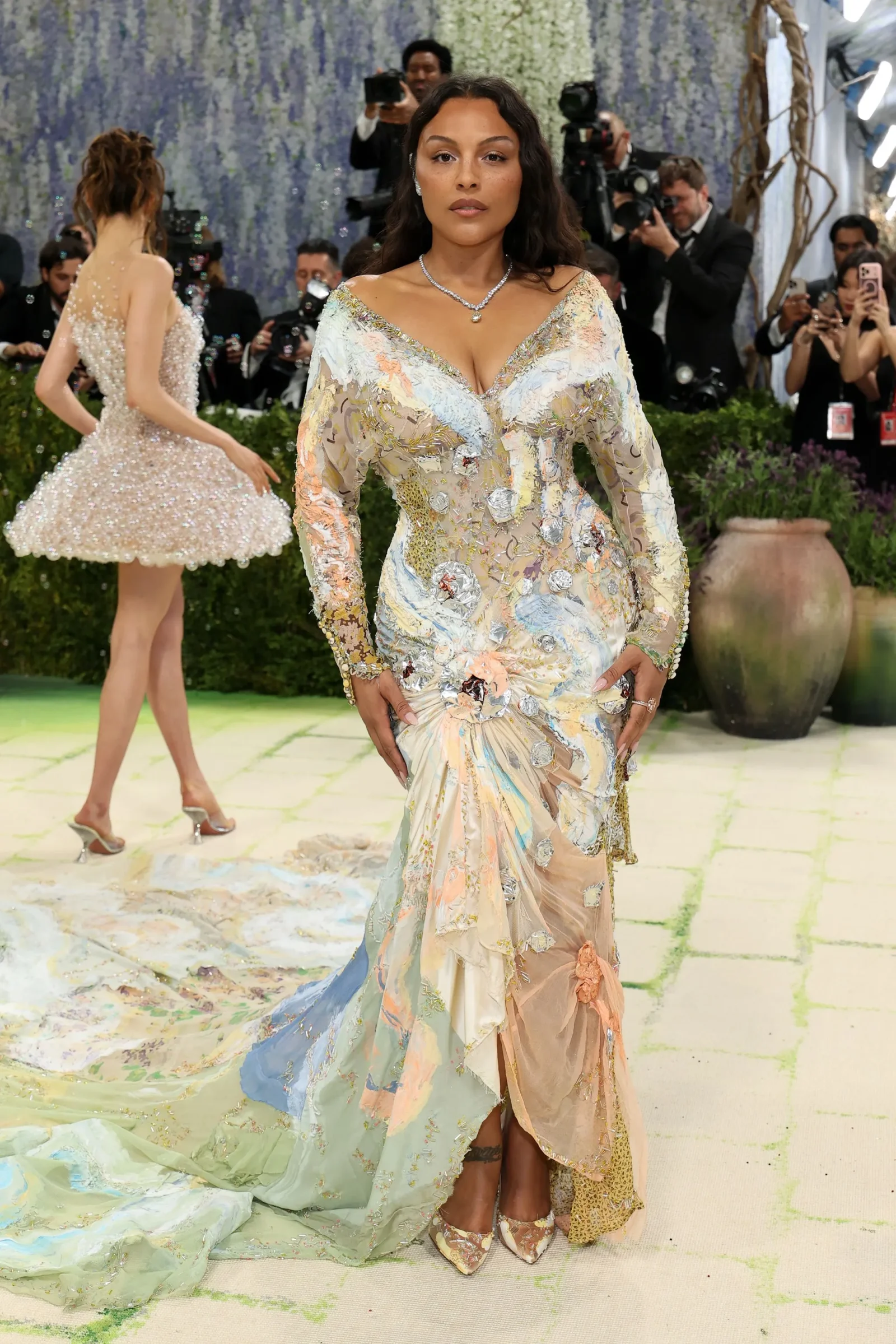 Center-stage woman wears a pastel beaded gown with a long, flowing train at a red-carpet gala; photographers and another gown-clad attendee in the background.