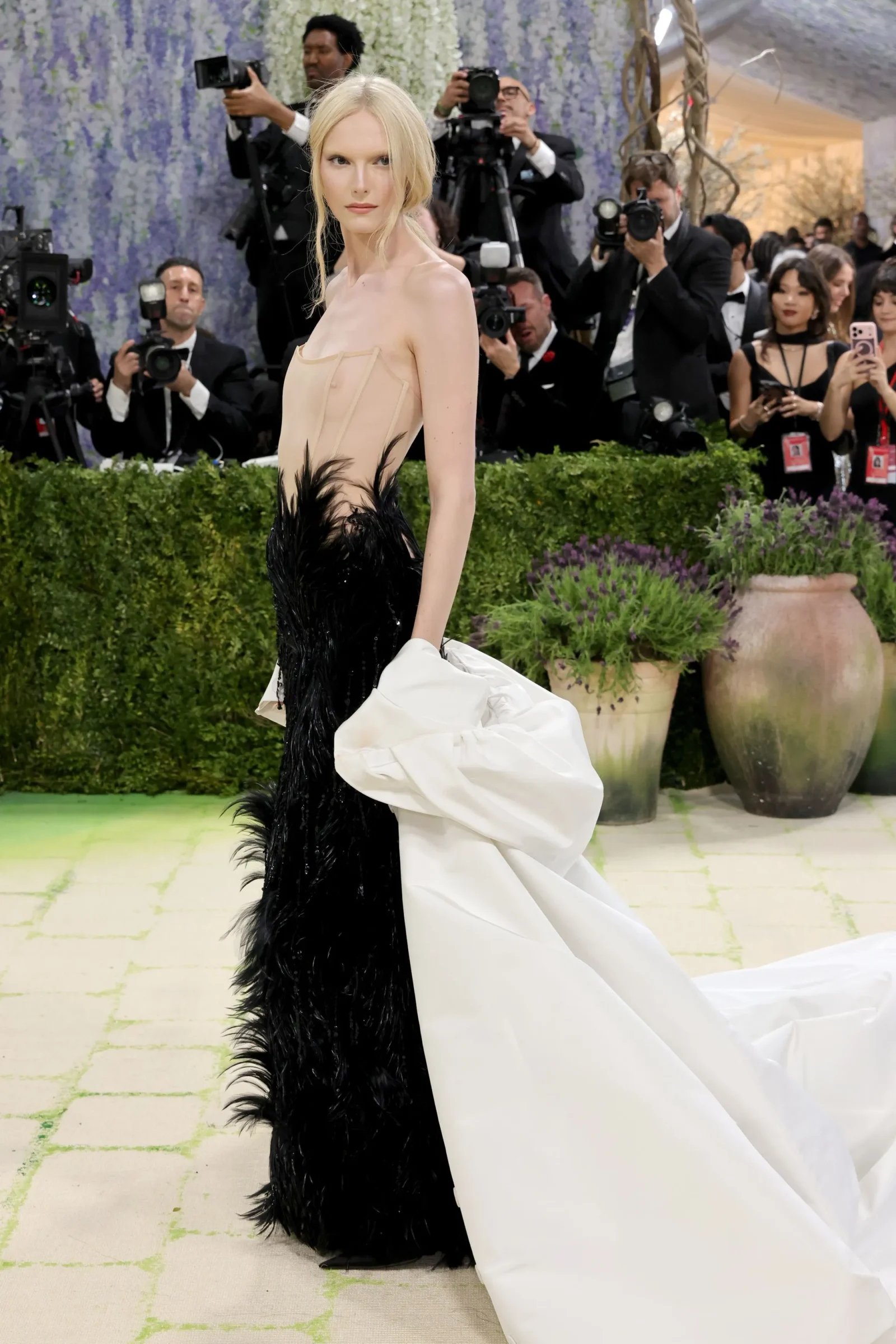 Blonde woman on a red carpet in a beige and black feathered gown, photographers behind her taking pictures.