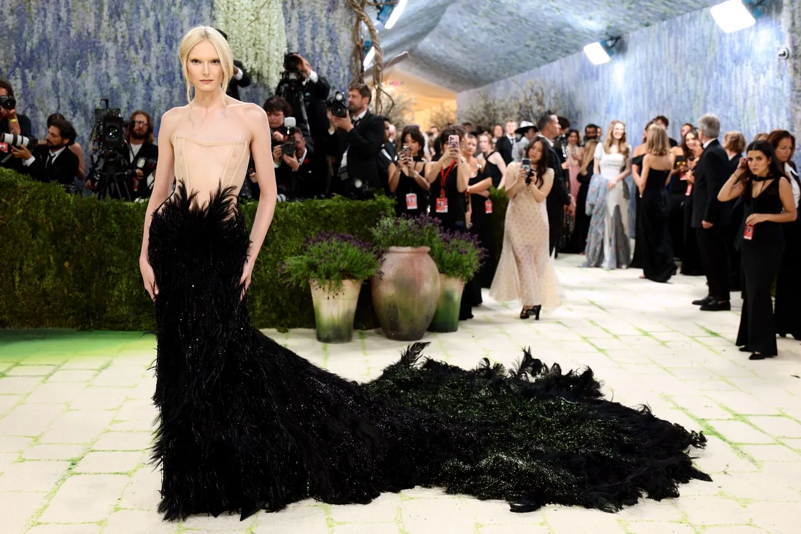Model wearing a strapless beige top with a dramatic black feathered train on a red carpet, photographers and guests in the background.