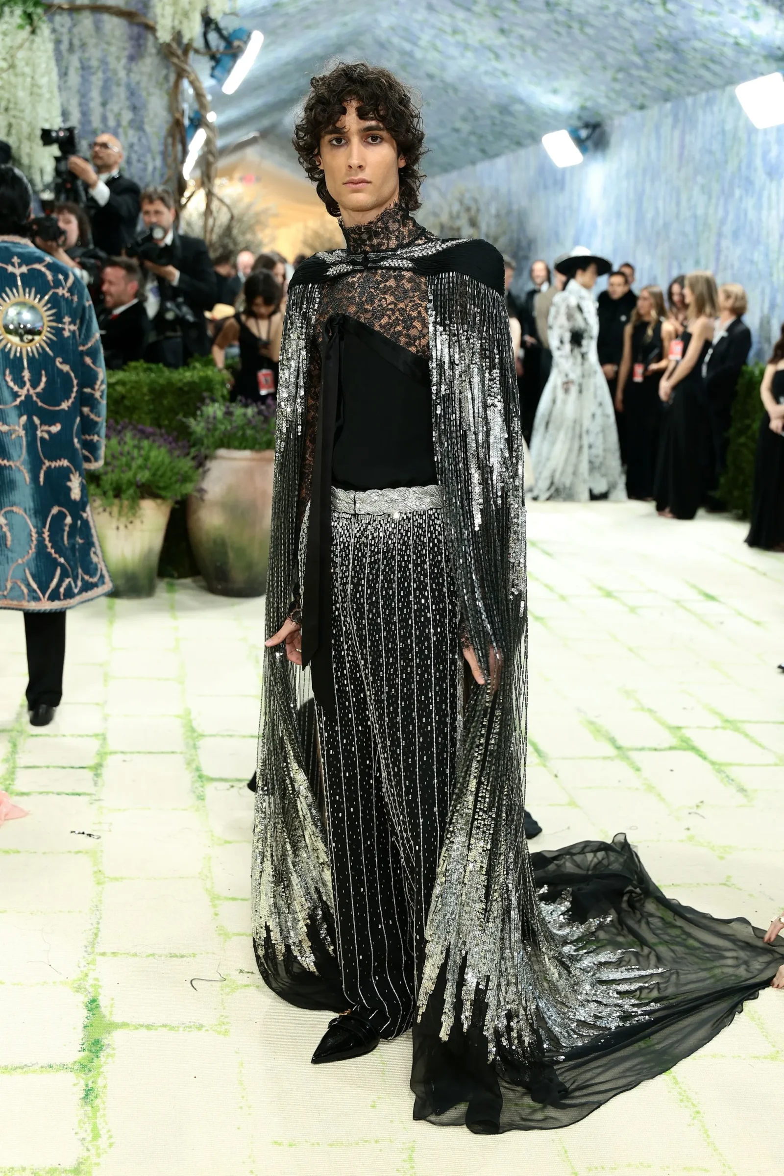 Model on a fashion runway wearing a black lace gown with long silver-fringe cape and a trailing train, photographers in the background.