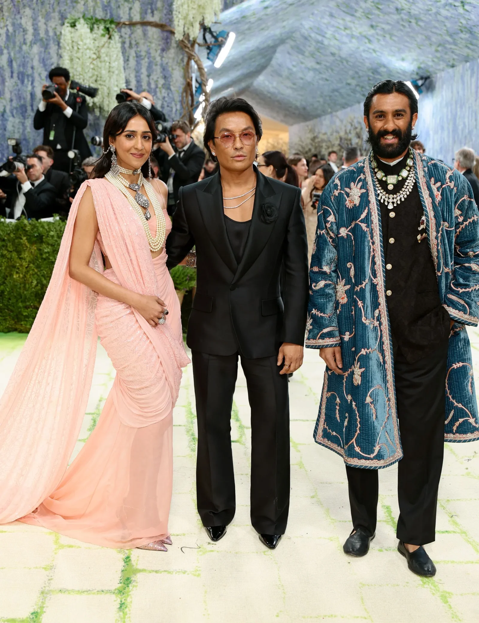 Three people pose on a fashion-event carpet: a woman in a pink saree, a man in a black suit with sunglasses, and a man in a blue embroidered coat, with photographers in the background.