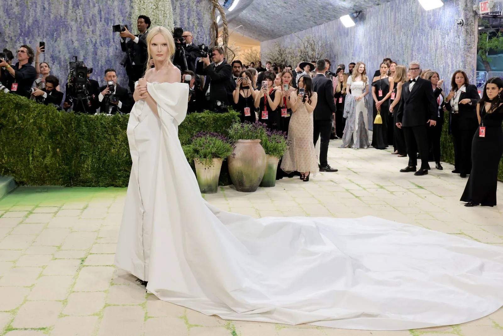 Woman in an off-shoulder white gown with a long train posing for paparazzi on a red carpet behind a hedge and photographers.
