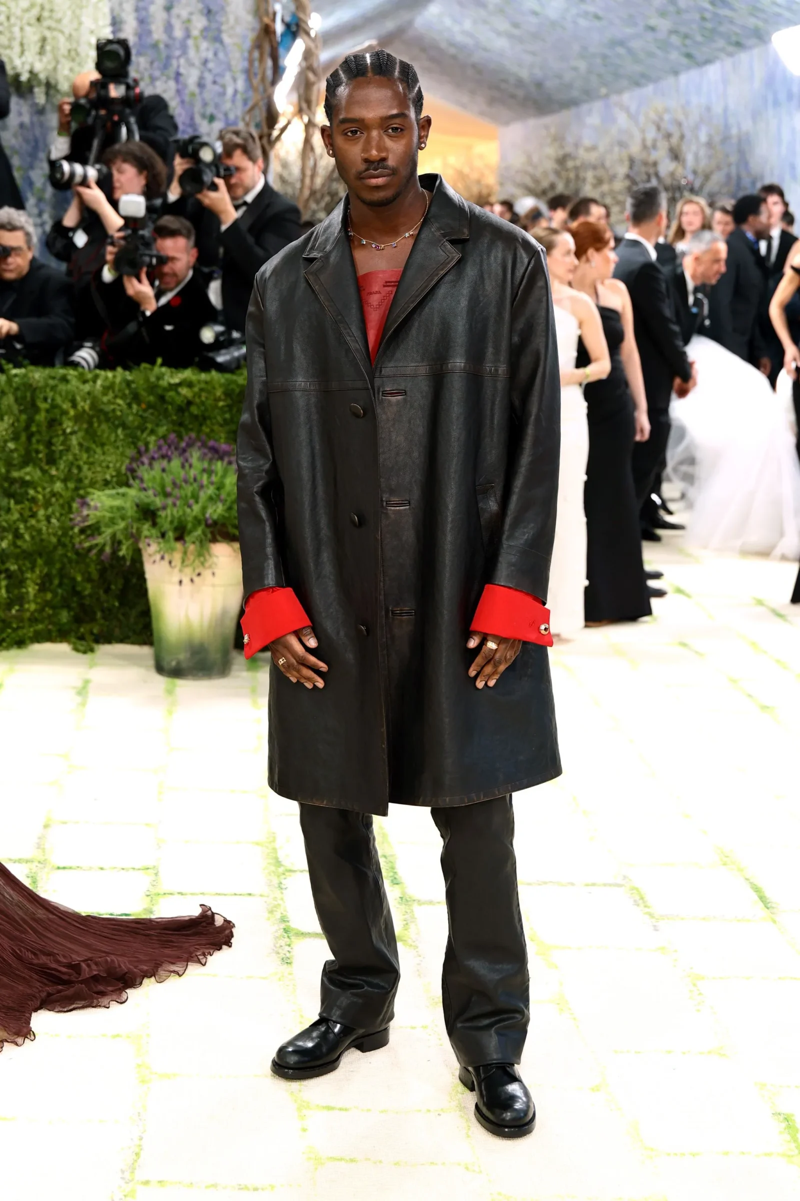 Man posing on a red carpet in a long black leather coat with red cuffs, photographers in the background