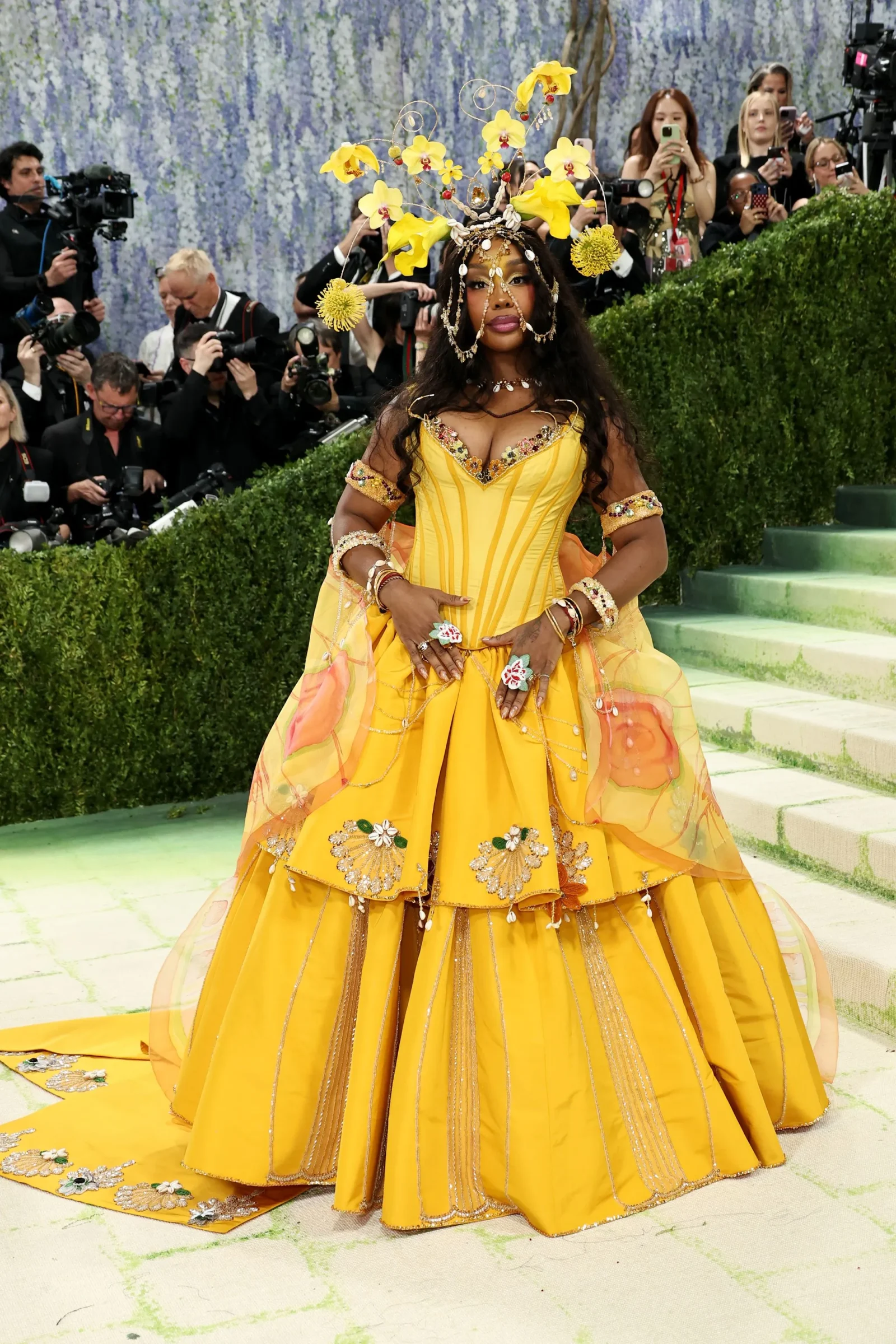 Woman in a bright yellow ball gown with a large floral headpiece posing on a red carpet, photographers in the background.