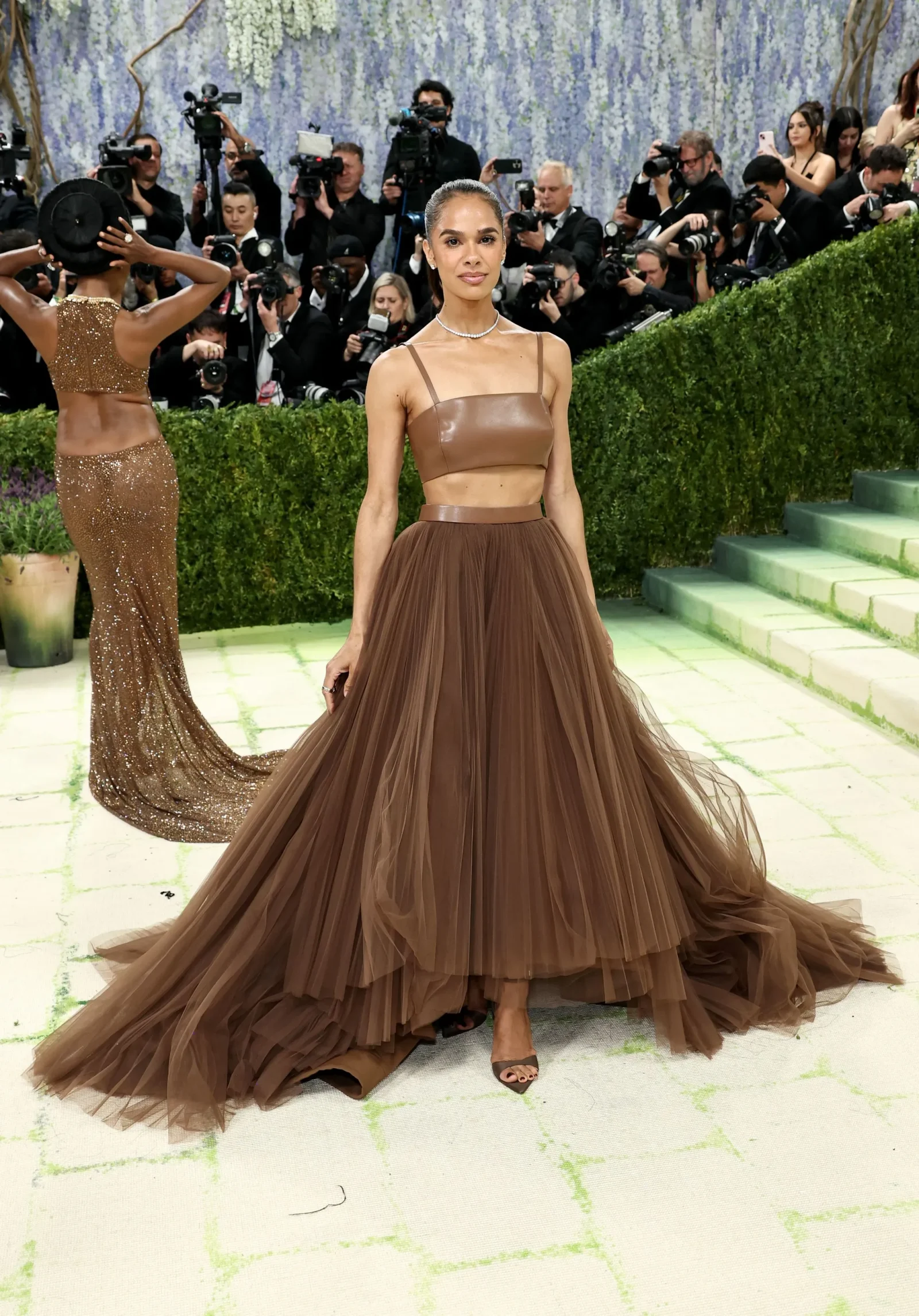 Woman on red carpet in a brown two‑piece gown with a voluminous tulle skirt, posing as photographers watch from behind a hedge backdrop.