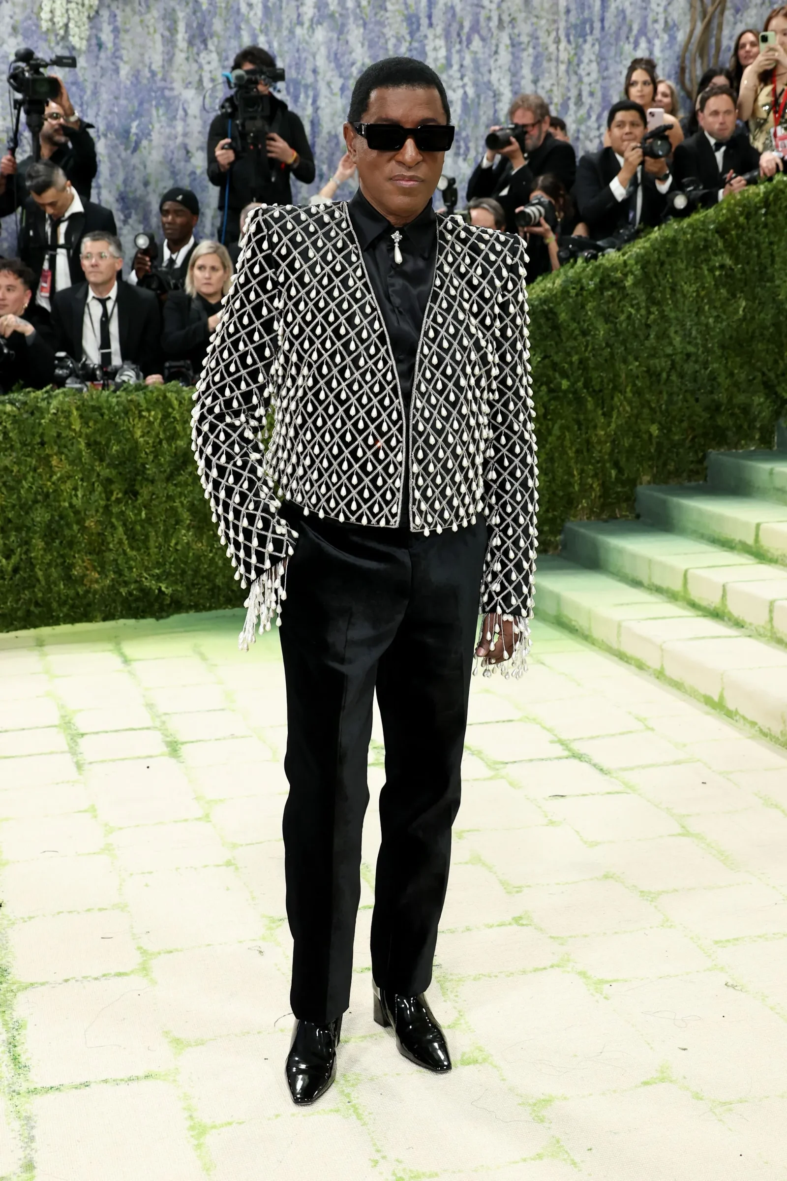 Person wearing a black outfit with a silver diamond-patterned jacket and sunglasses at a red carpet event.