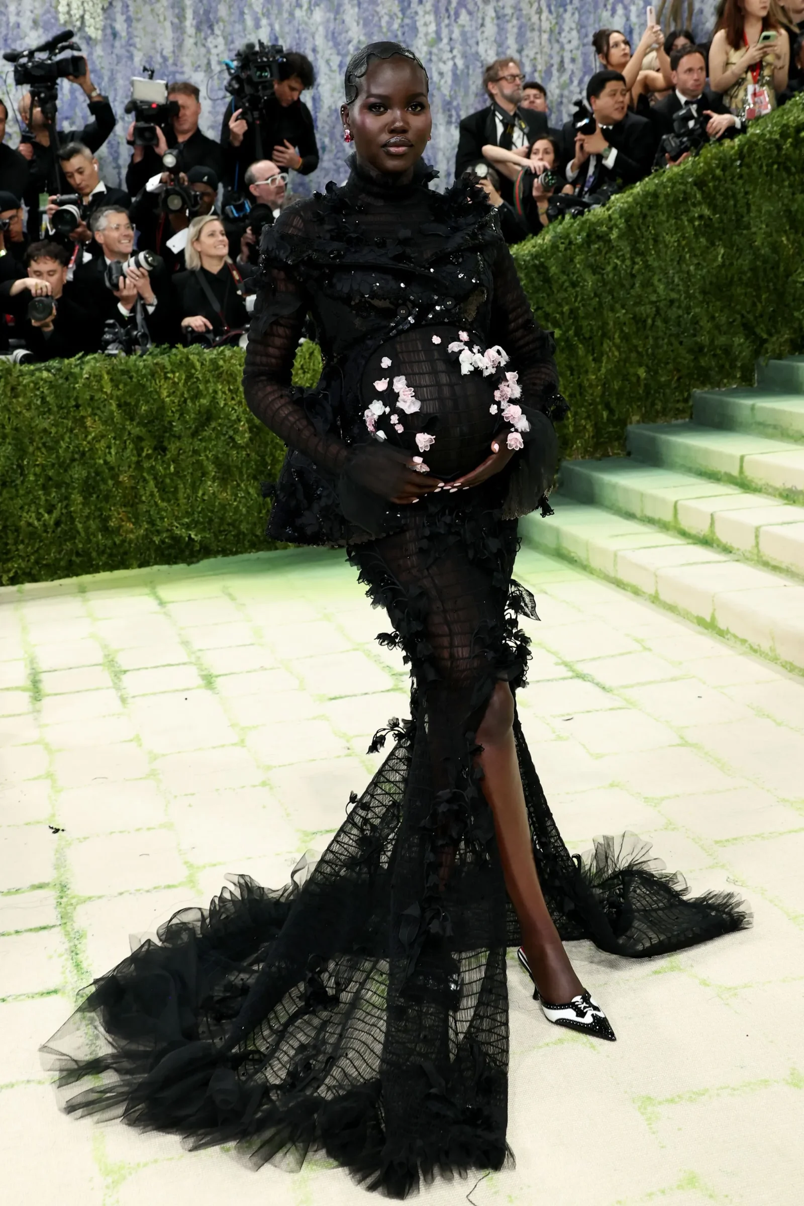 Pregnant woman in a black floral gown posing on a red carpet with photographers behind her.