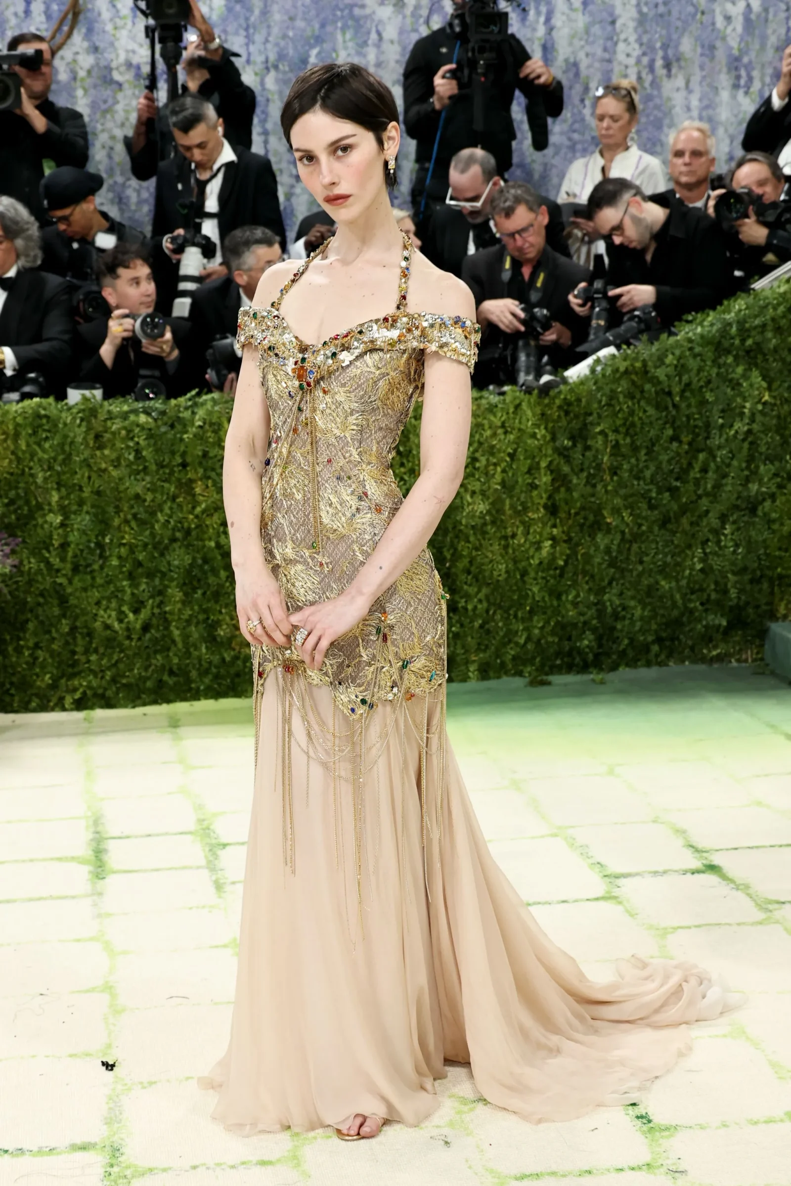 Model in an ornate gold beaded evening gown posing while photographers circle around her on a fashion show set.