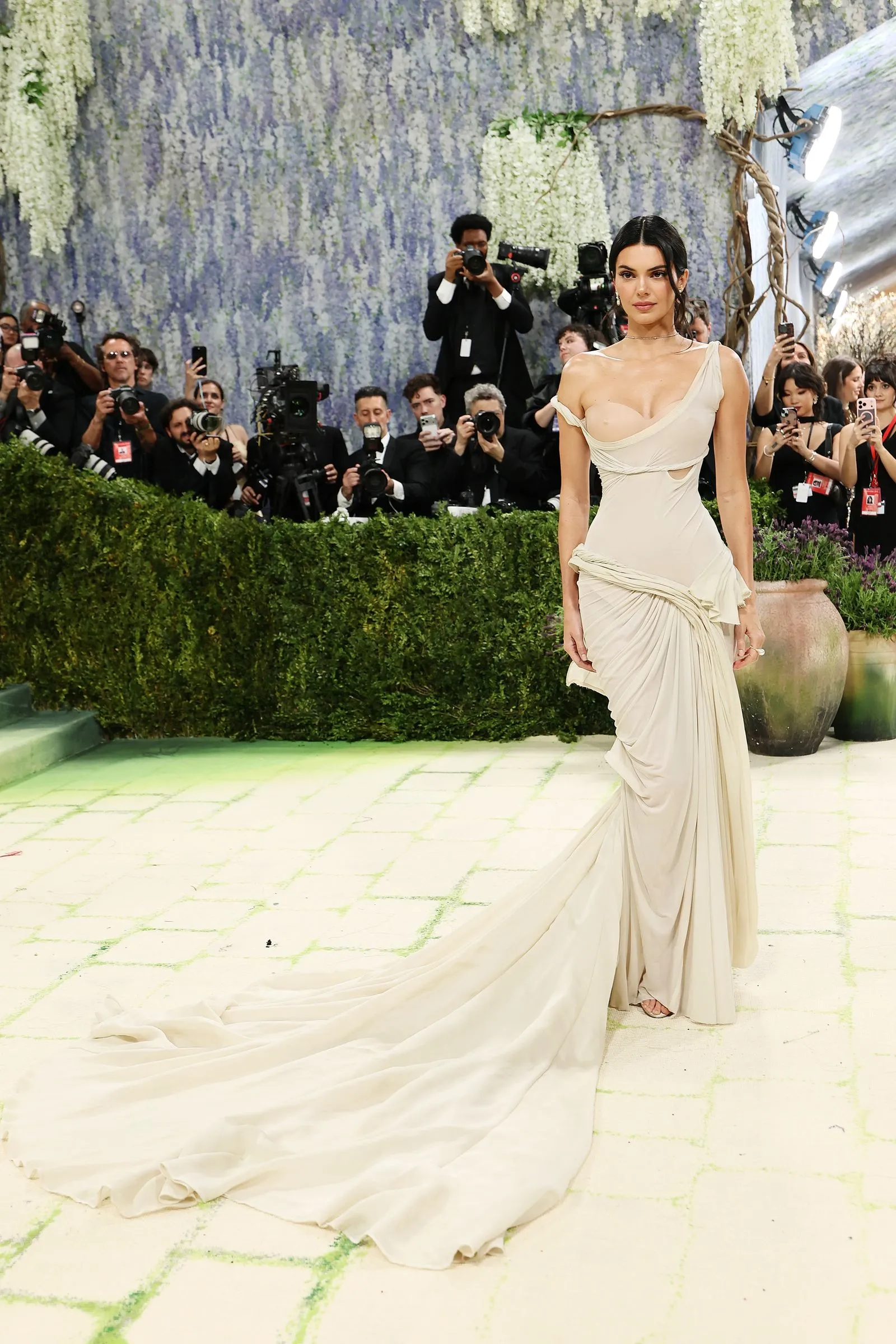 Woman in an elegant cream gown with a long train posing for cameras at a glamorous event, photographers behind her.