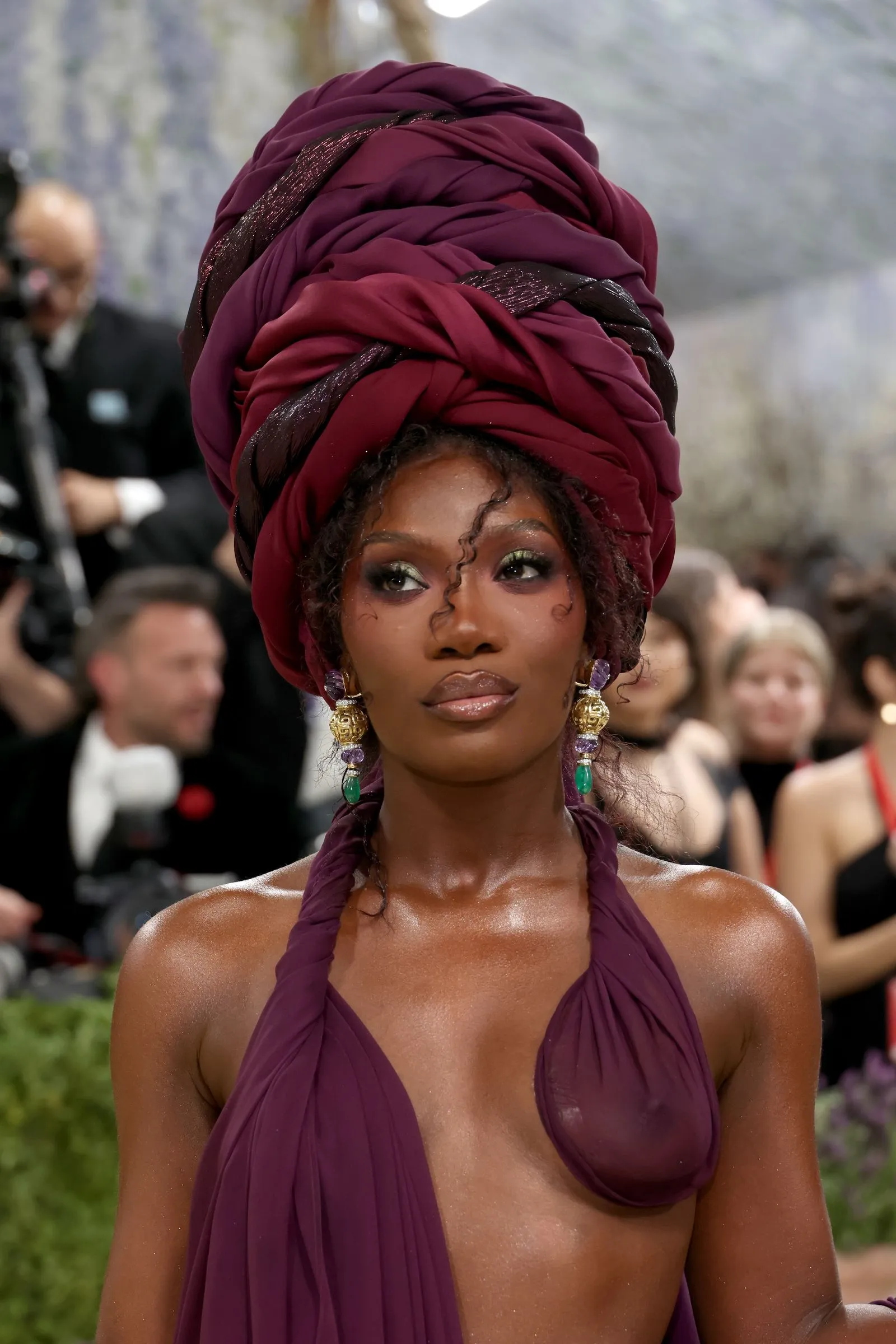 Woman wearing a voluminous burgundy head wrap and matching dress, gold jewelry, on a red carpet event.