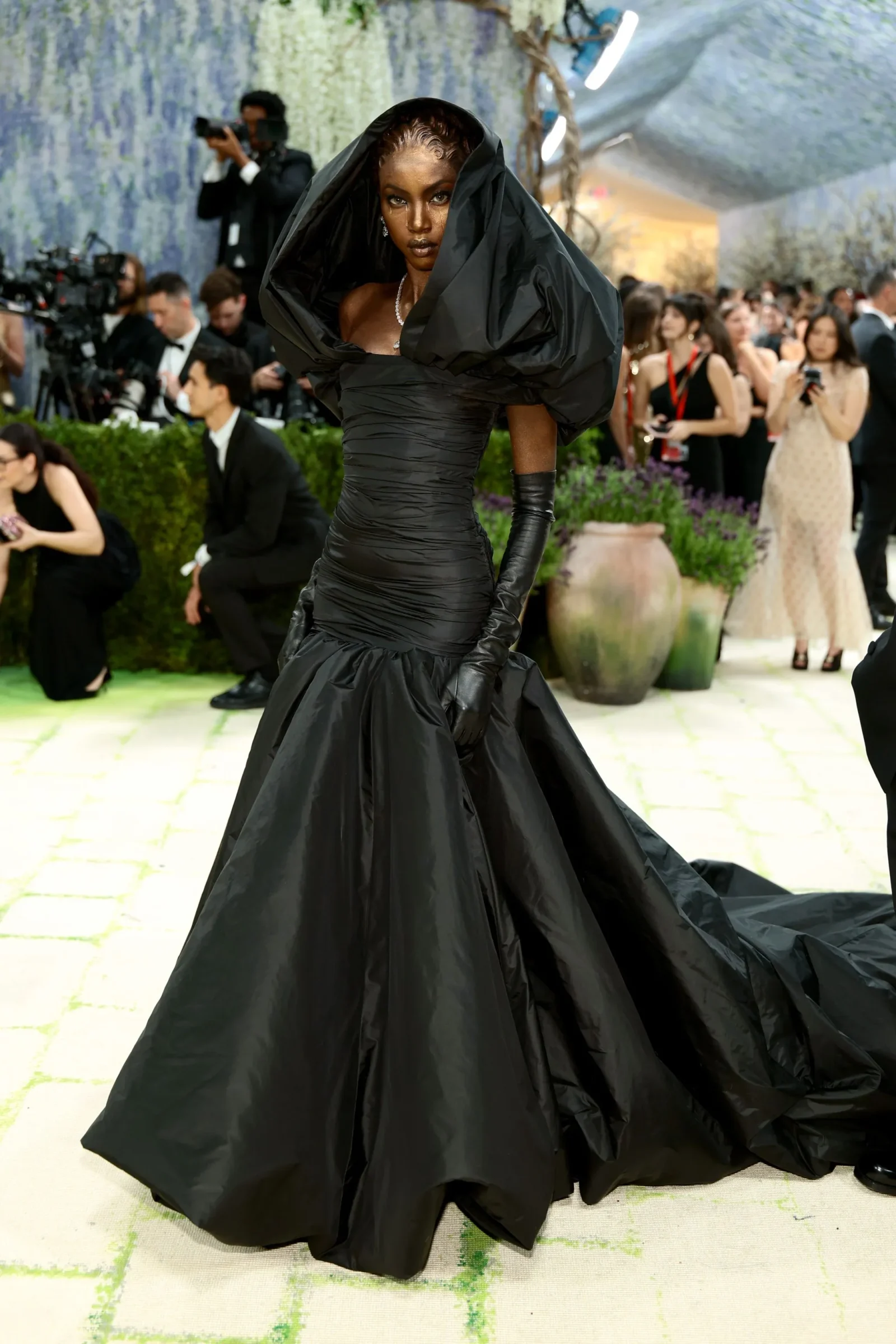 Model wearing a dramatic black gown with a voluminous draped hood on a red carpet, photographers in the background