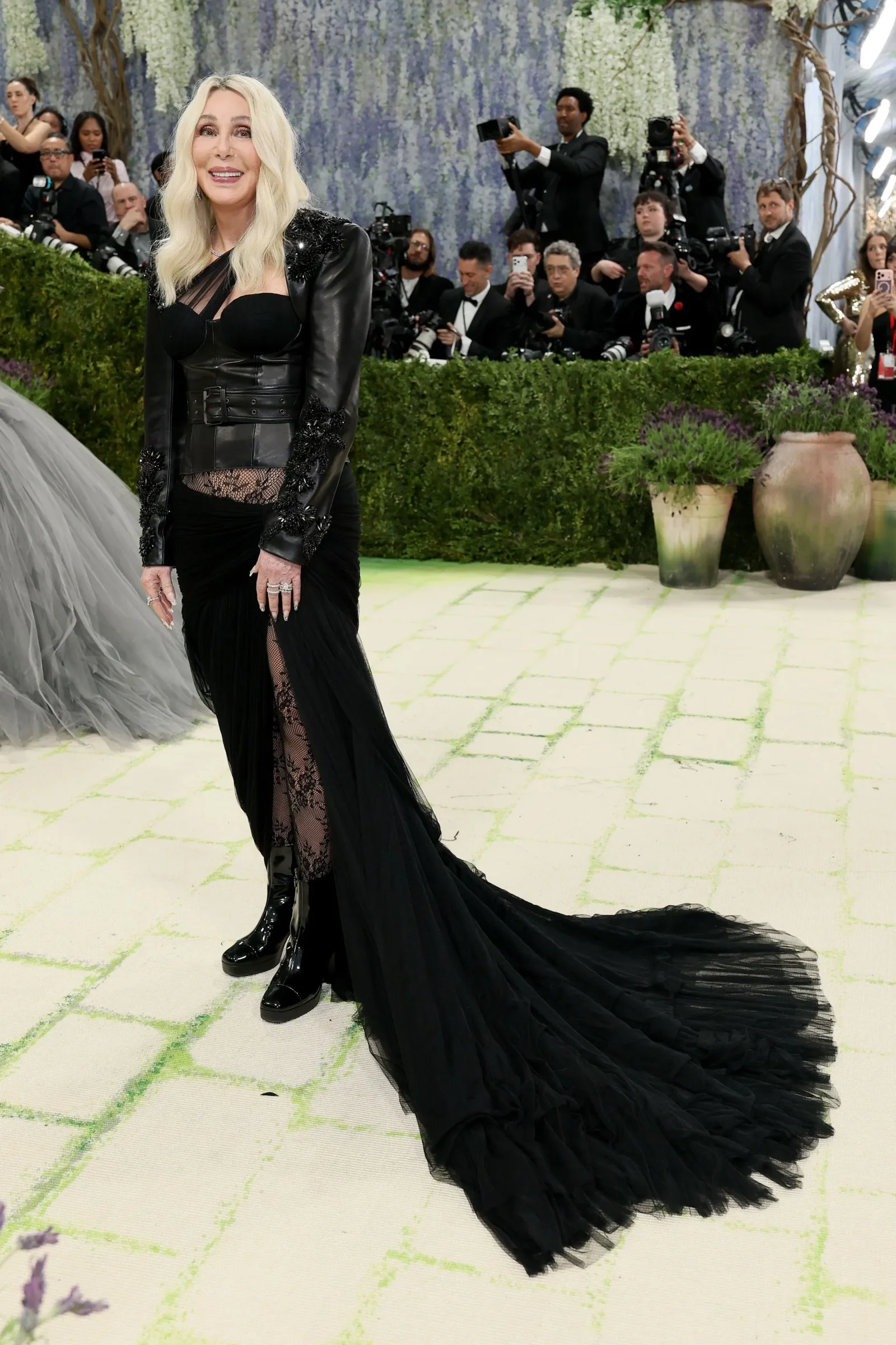 Blonde woman in a black leather gown with lace detailing posing on a pale tiled carpet as photographers shoot in the background.