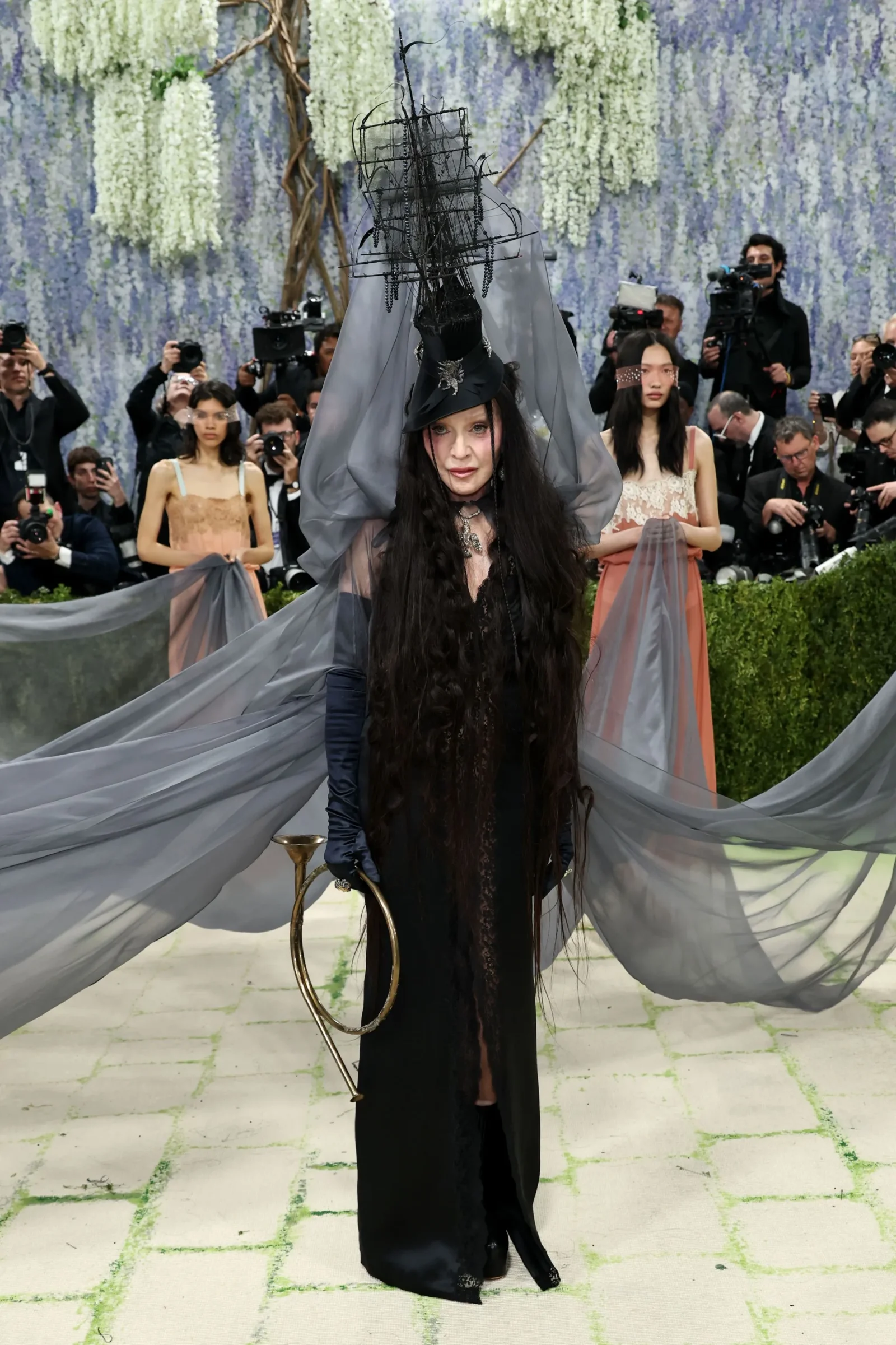Model in an extravagant black gown with a tall, sculptural headpiece and sheer gray cape, posing amid photographers at a flower-filled runway.