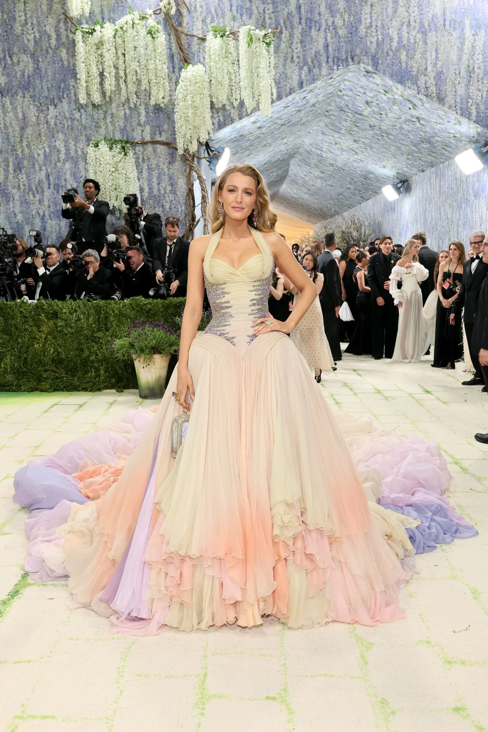 A woman in a flowing pastel pink gown posing on a green-and-white patterned runway as photographers line the backdrop behind her.