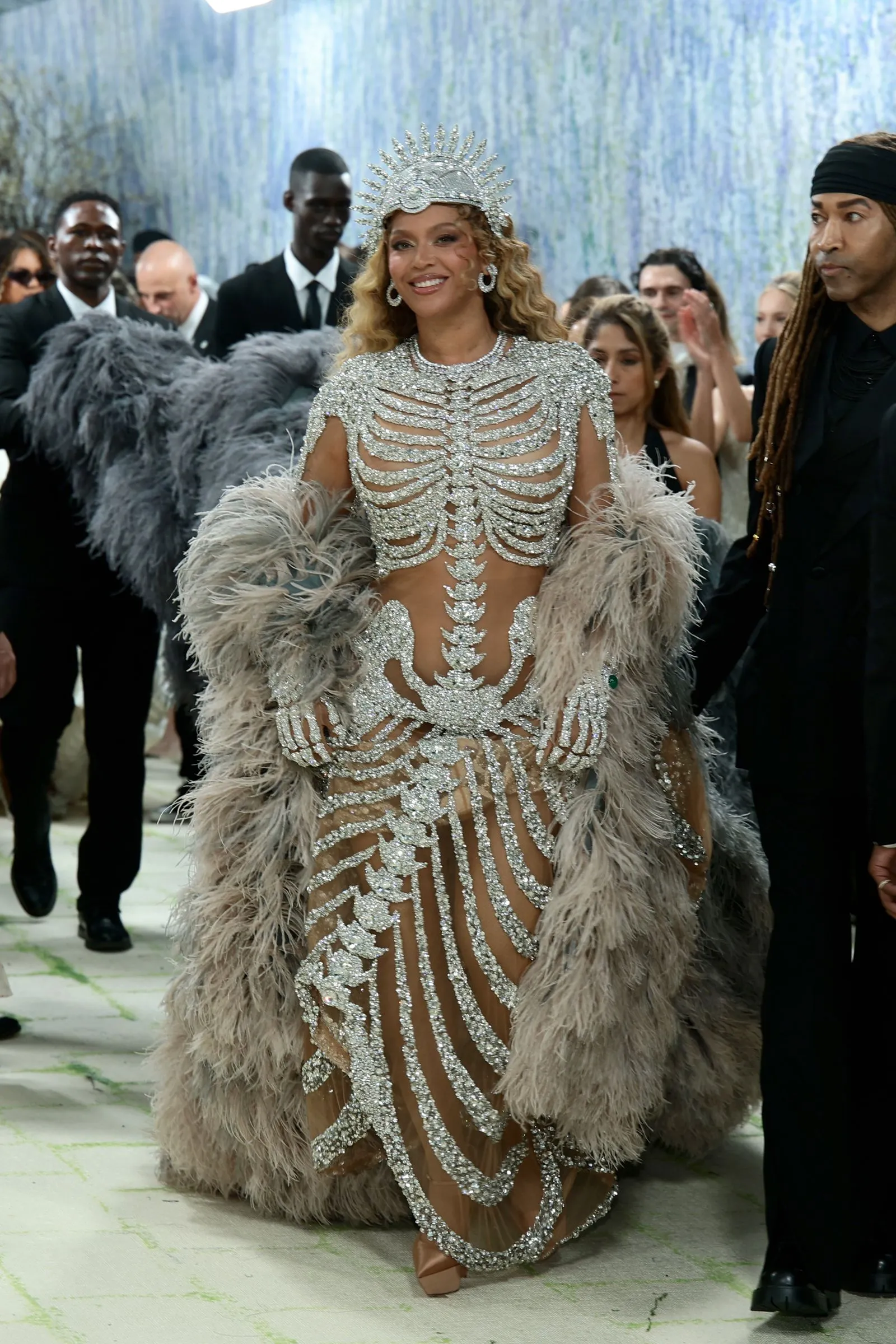 Model walking a runway in a silver, gem-encrusted gown with cutouts and feathered cape at a fashion show.