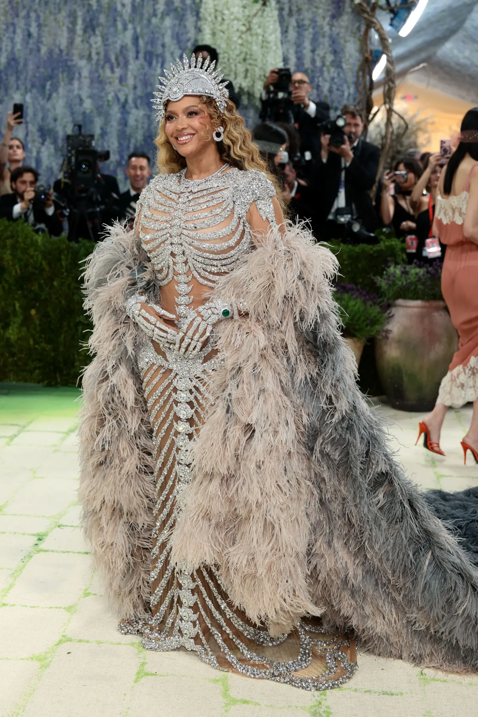 A glamorous woman on a red carpet wearing a metallic, bejeweled bodysuit and a large feathered cape, with a jeweled crown and statement earrings; photographers in the background.