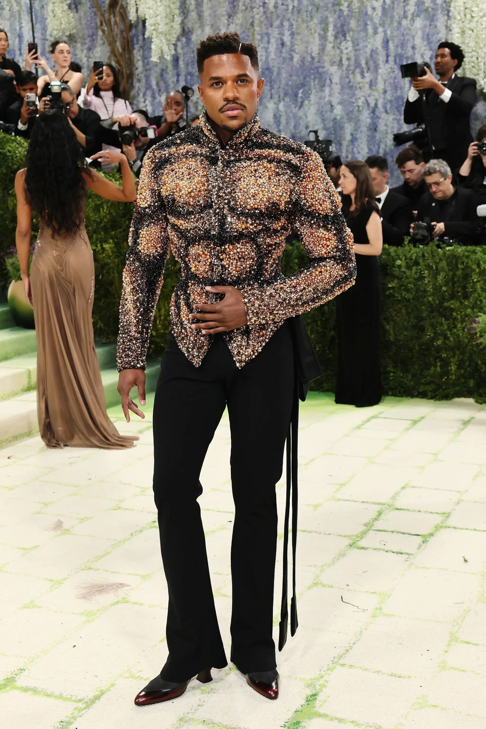 Man posing on a green carpet in a black-and-gold gem-studded jacket at a formal event, photographers in the background.