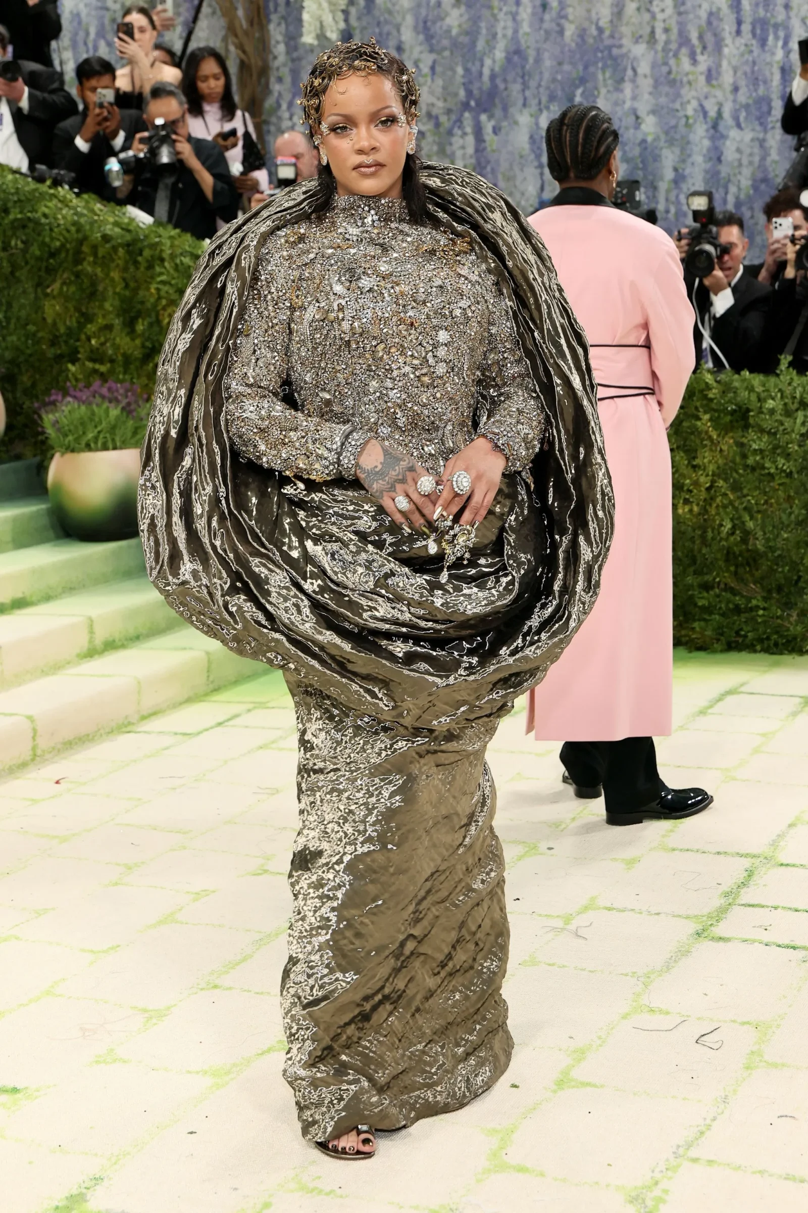 Rihanna in a heavily jeweled, metallic gown with an voluminous patterned cape, standing on a green carpet at a high-profile event while photographers look on