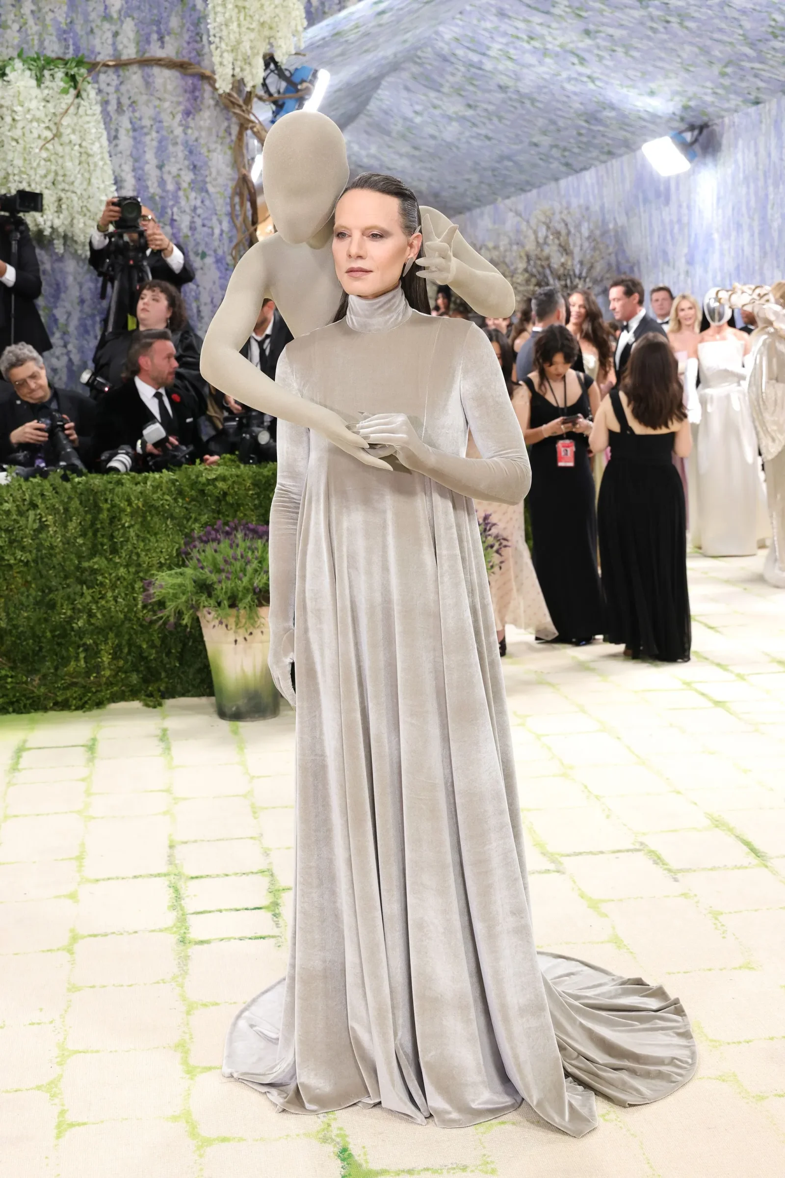 Model in a silver, floor-length gown with a large sculptural headpiece and arm extensions on a fashion show runway, photographers in the background.