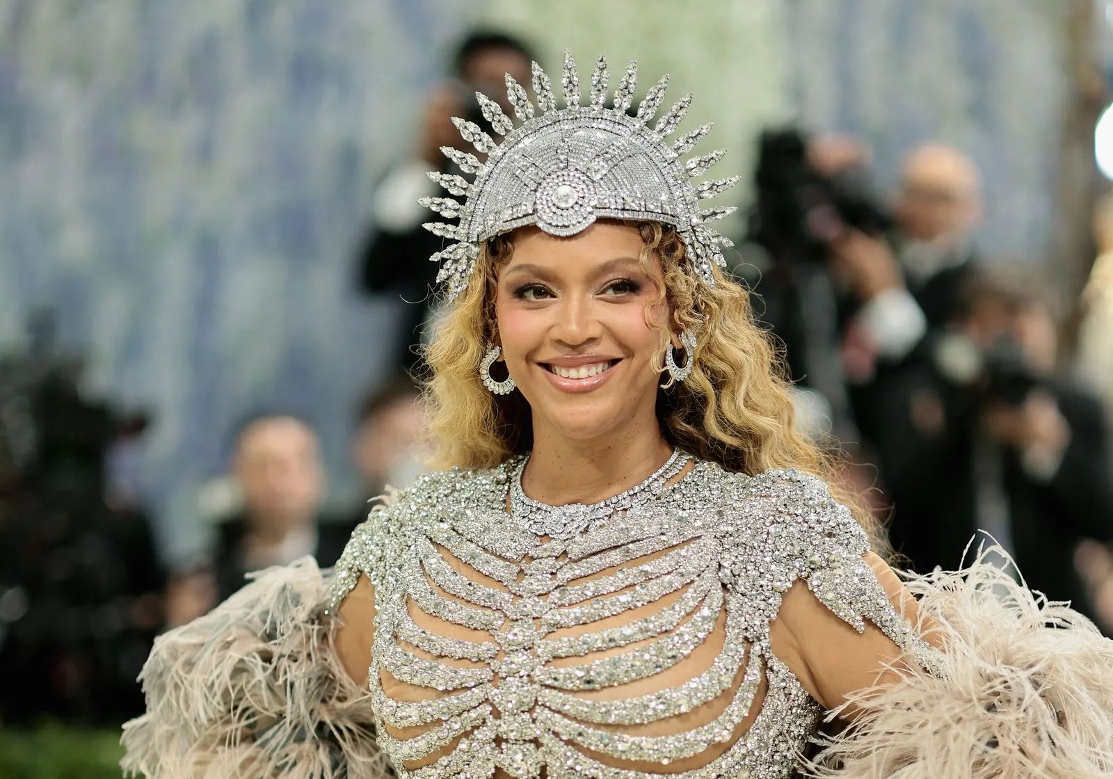 Smiling woman in a silver jewel-encrusted gown with a sunburst headpiece and feathered sleeves on the red carpet.