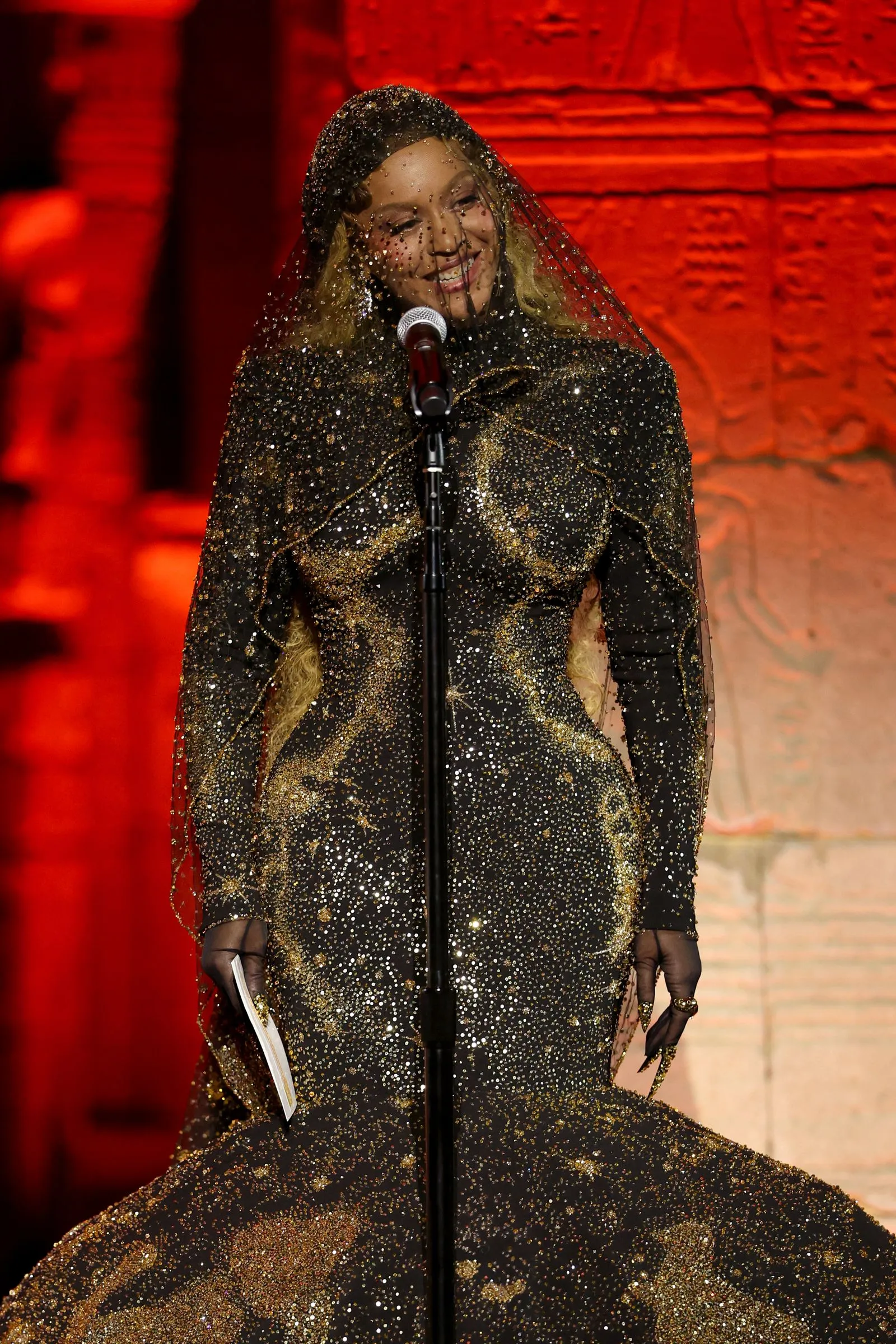 Female performer in a glittery black and gold gown with a sheer veil, singing into a microphone on a red-lit stage.