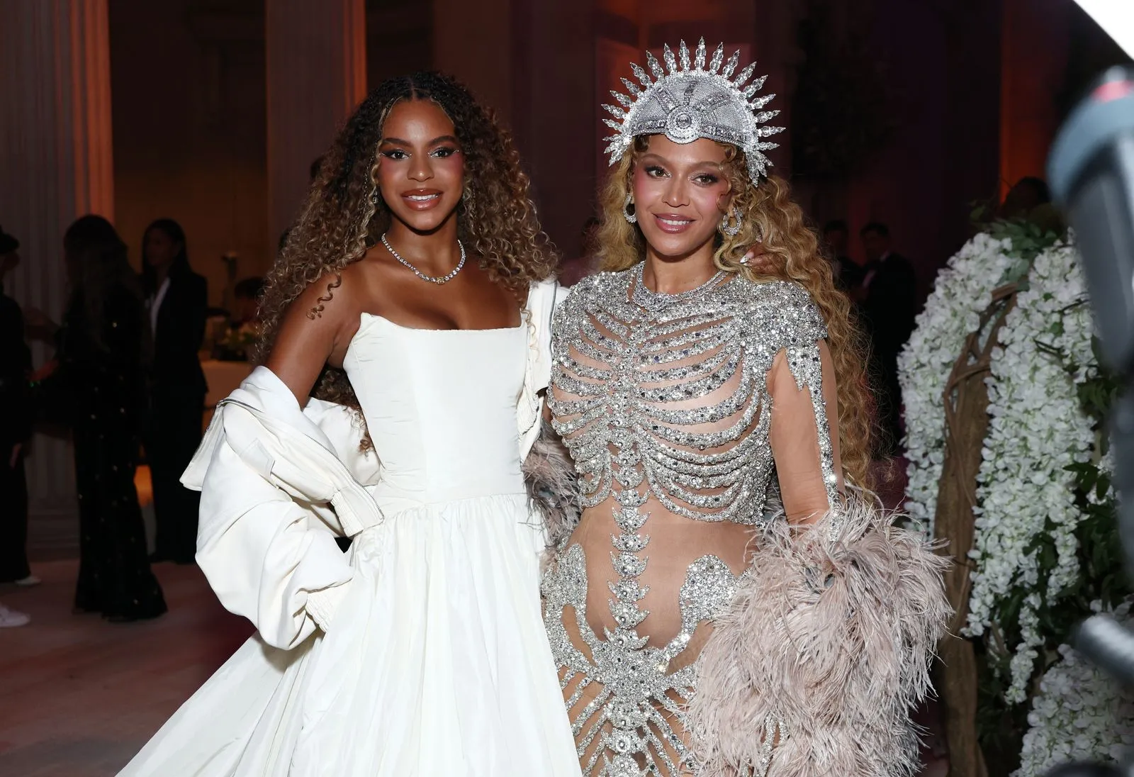 Two women in formal gowns pose together at a gala: one in a white off‑shoulder dress, the other in a silver jeweled outfit with a crown and feathered train.
