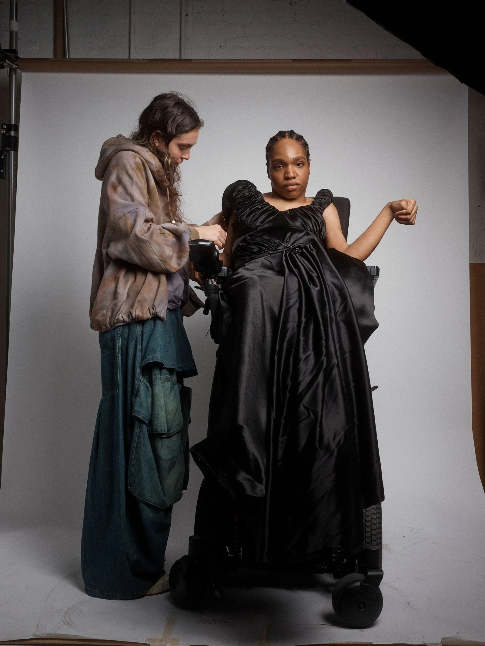 A stylist adjusting a black satin gown on a person in a wheelchair inside a photography studio.