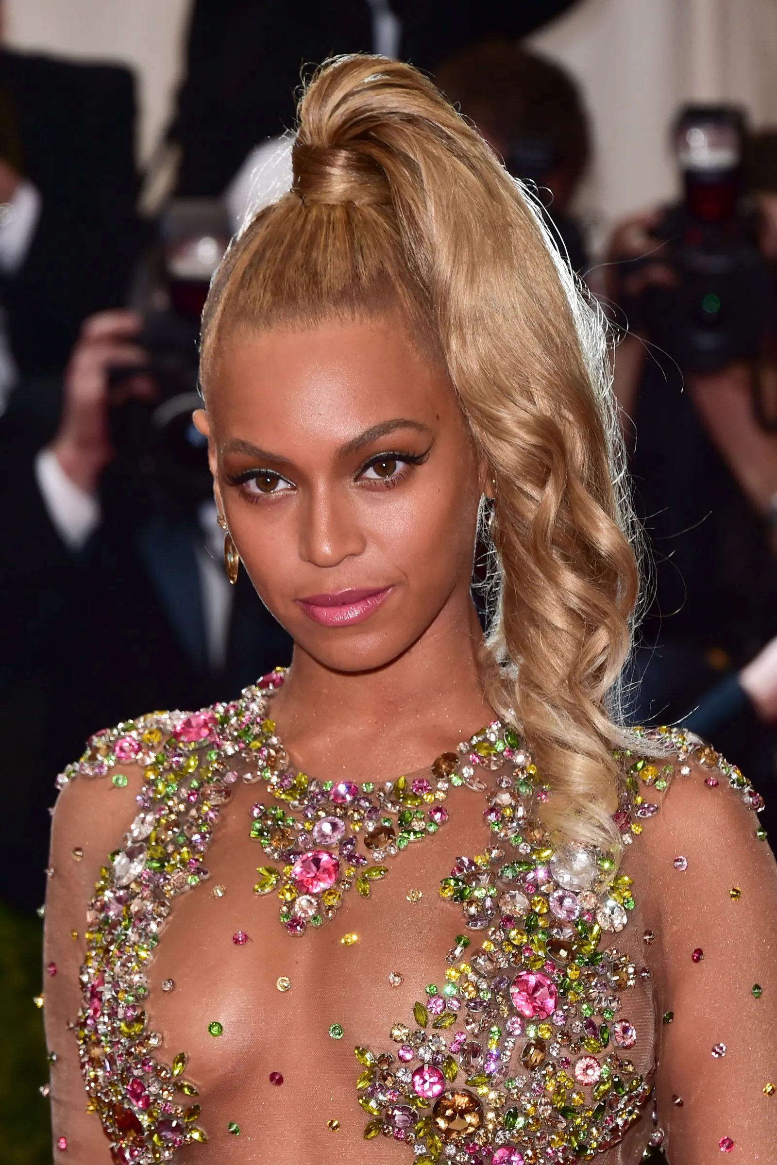 Woman with a high blonde ponytail and a jewel-covered sheer gown on the red carpet, photographers in the background.