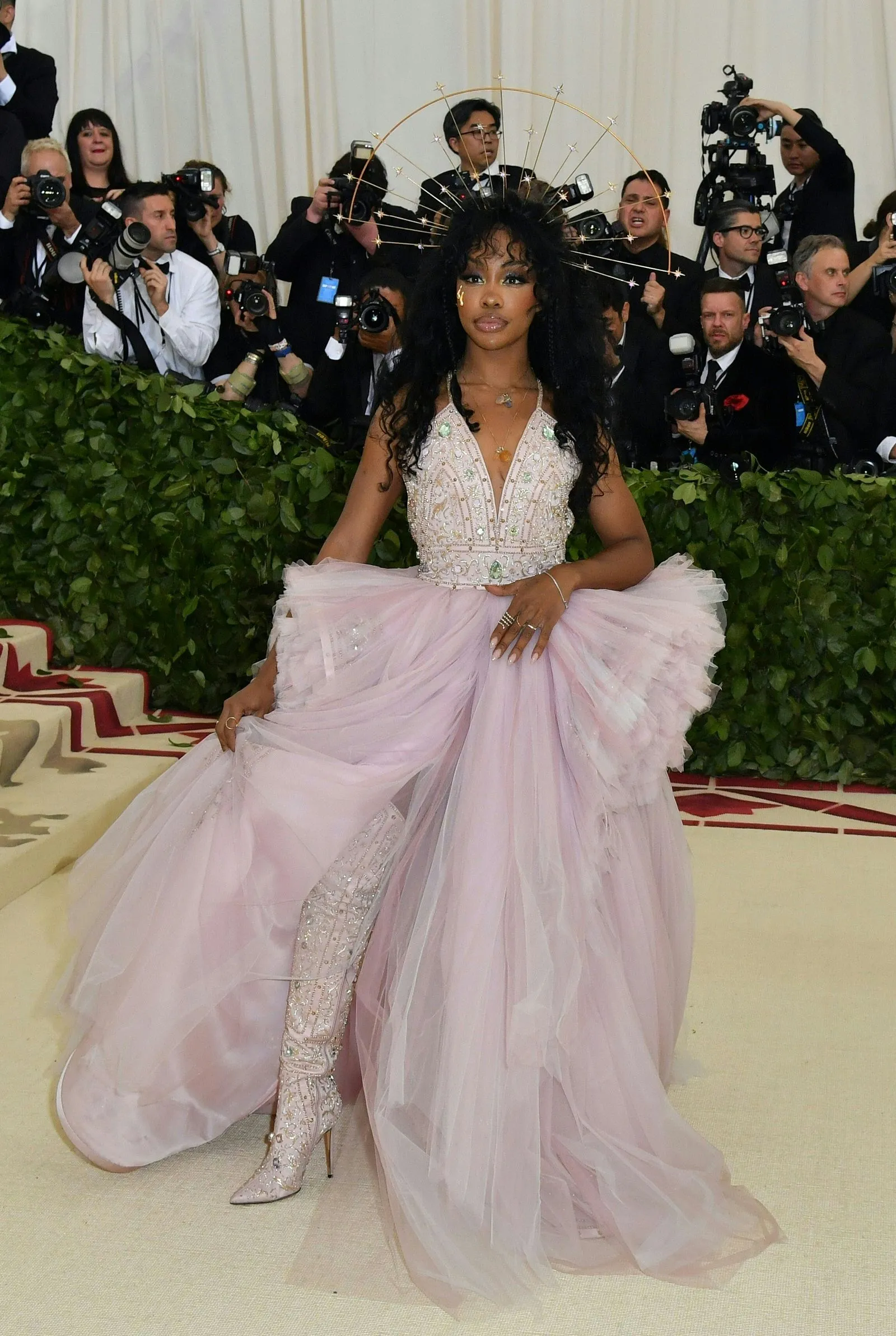 Woman wearing a pale pink, beaded ball gown with a voluminous tulle skirt poses on a red carpet as photographers crowd around in the background, wearing a starburst halo headpiece.